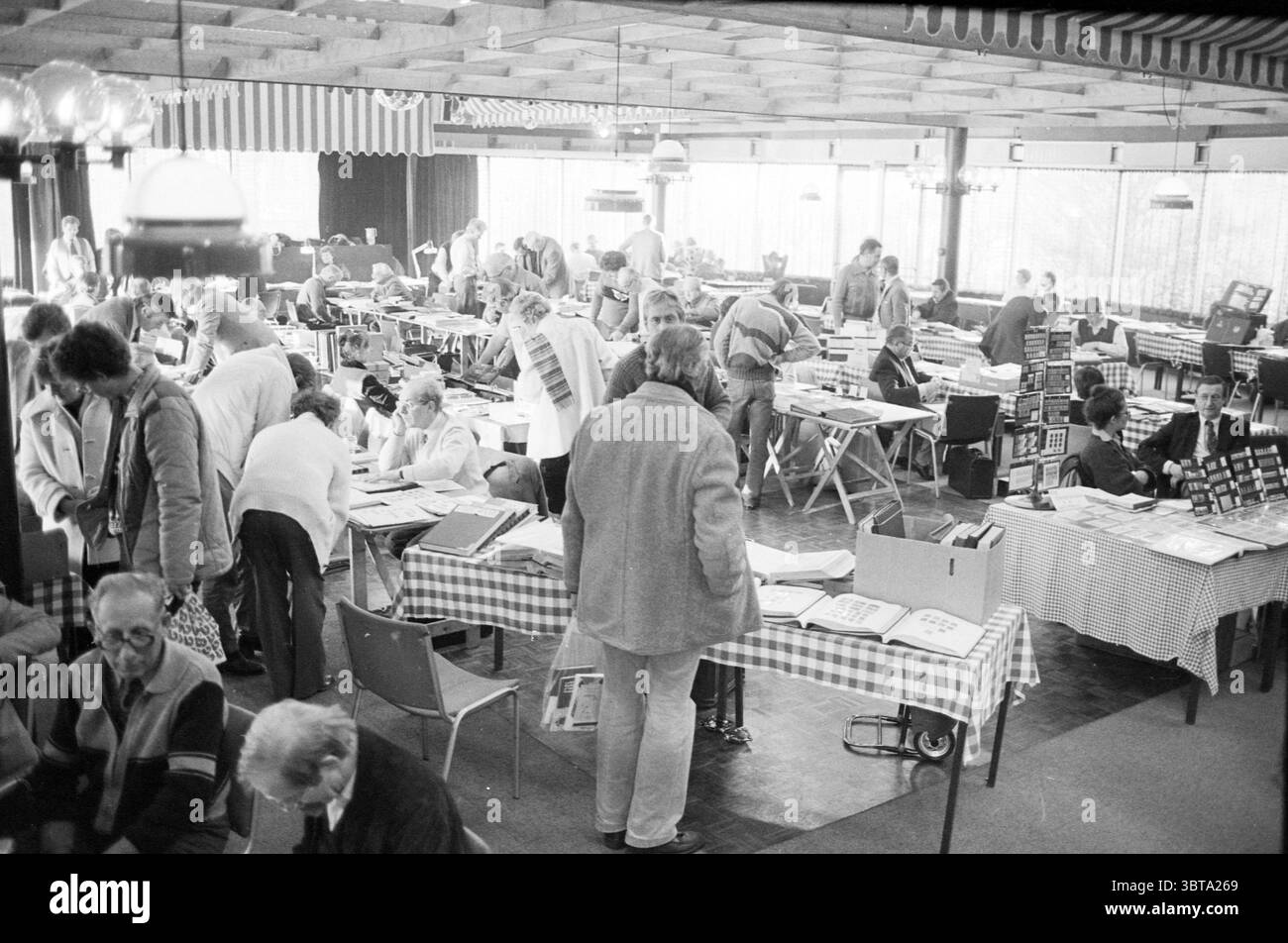 Fiera di francobolli e monete, Whizgle News, Dutch Desk, Paesi Bassi, 1950 - 2000 il 12-1983. L'immagine include questi argomenti. La scena mostra un vivace spazio interno pieno di numerosi tavoli e persone impegnate in varie attività. Il pavimento è in legno, a complemento dell'atmosfera calda e invitante creata da una miscela di illuminazione naturale e artificiale. Una varietà di tavoli, avvolti in panni a scacchi, sono sparsi in tutta la stanza, con un assortimento di oggetti come libri, fotografie e opere esposte. Persone di età diverse, alcune sedute mentre altre si alzano o spostano abou Foto Stock