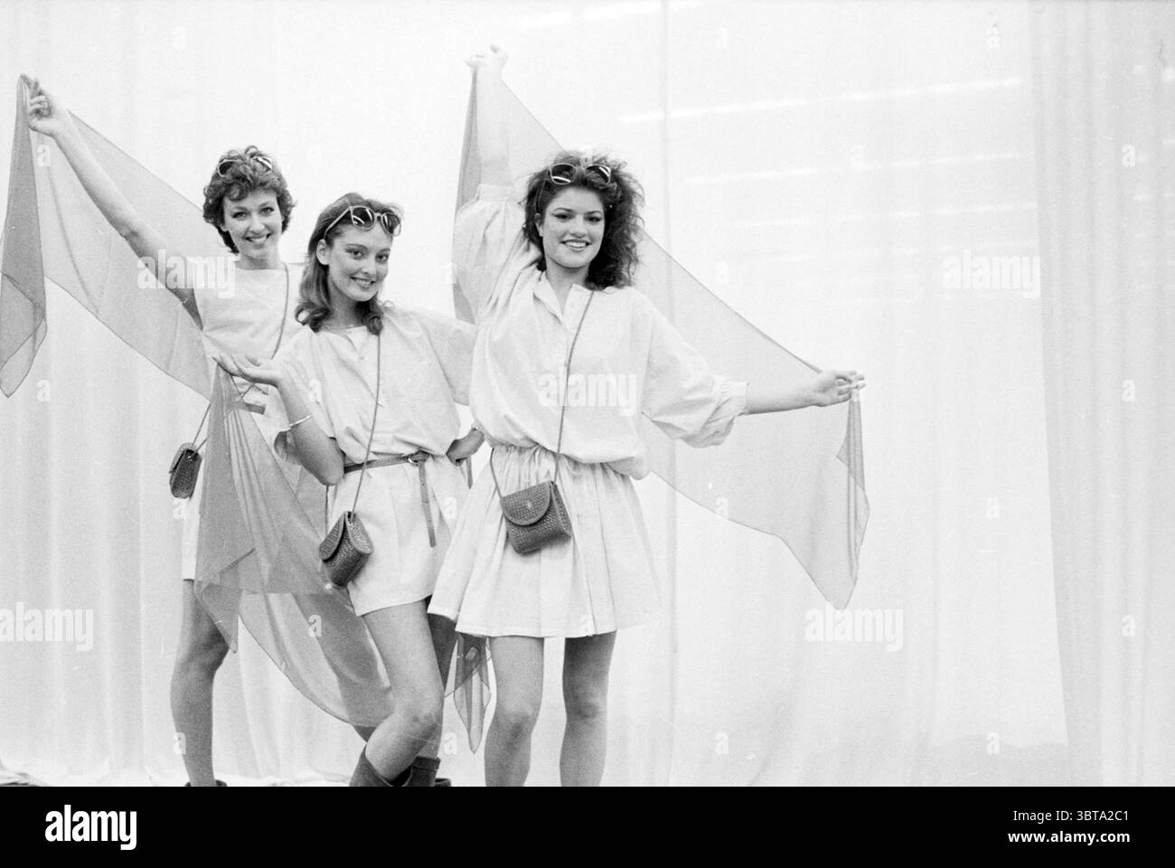 Tendenze della moda di primavera Amsterdam Fashion Amsterdam Paesi Bassi, Whizgle News, Dutch Desk, Paesi Bassi, 1950 - 2000 il 19-04-1983. L'immagine mostra questi argomenti. Nella scena, tre modelli sono posizionati su uno sfondo morbido e leggero che crea un'atmosfera ariosa ed eterea. Sono indossati con indumenti bianchi e aderenti che donano un senso di eleganza casual. Gli outfit sono completati da tessuti morbidi e trasparenti in tonalità delicate che catturano la luce, suggerendo movimento e grazia come sono tenuti dai modelli. Il primo modello è a sinistra, con un'espressione giocosa, lui Foto Stock