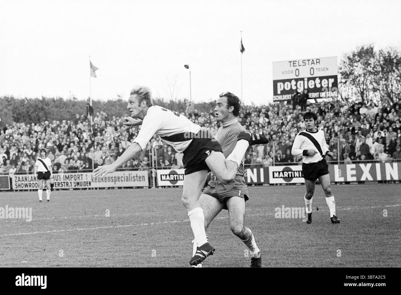 Telstar - FC Den Bosch Football Telstar, Whizgle News, Dutch Desk, Paesi Bassi, 1950 - 2000 il 14-11-1971. L'immagine mostra questi argomenti. La scena cattura un momento intenso su un campo da calcio, pieno di azione ed energia. In primo piano, due atleti sono bloccati in una lotta dinamica. Un giocatore, indossando una maglia bianca con bordo nero, sta saltando in aria, mostrando un senso di urgenza e atleticità, mentre l'altro, con l'abito di un portiere, sta raggiungendo, forse tentando di intercettare la figura aerea o reagire a un gioco. Intorno a loro, una folla diversificata riempie le tribune, s Foto Stock