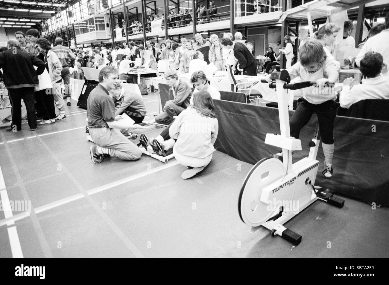 Veronica Fitrace Kennemer Sports Hall settimana dello sport e dello sport, Whizgle News, Dutch Desk, Paesi Bassi, 1950 - 2000 il 13-03-1988. Questi sono gli argomenti nell'immagine. La scena è piena di attività in uno spazio interno pieno di persone impegnate in varie attività fisiche. Il pavimento è contrassegnato da linee che indicano una palestra o una palestra polifunzionale. A sinistra, un gruppo di persone è concentrato su una macchina per esercizi seduti. Due persone stanno regolando l'attrezzatura mentre un'altra sta dimostrando il suo utilizzo, mostrando uno spirito di lavoro di squadra e incoraggiamento. Nelle vicinanze, un ragazzo sta pedalando Foto Stock