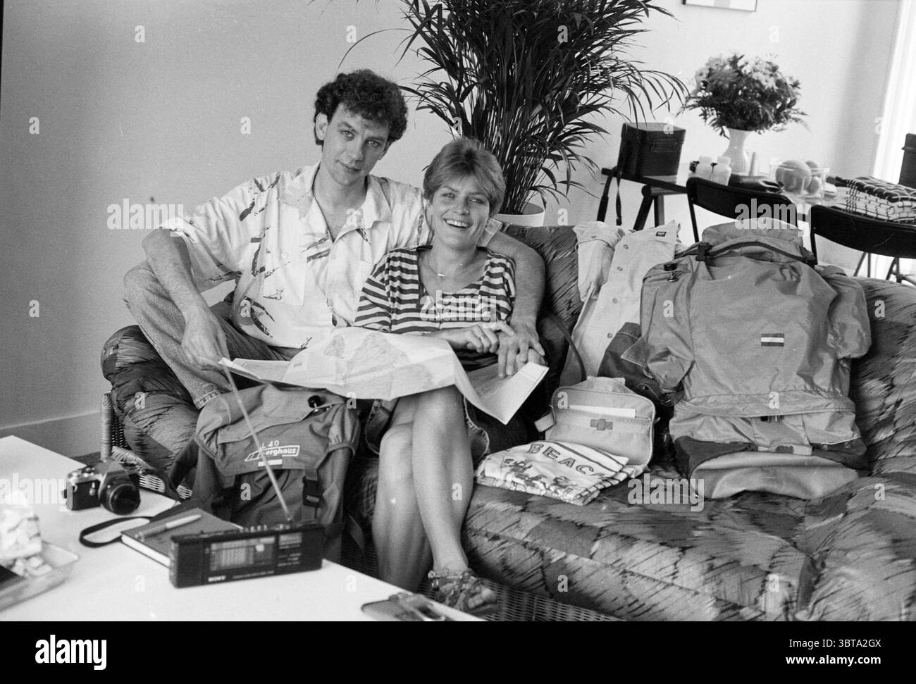 Tineke Ruiter and Wouter van Egmond World Travelers Travel, Whizgle News, Dutch Desk, Paesi Bassi, 1950 - 2000 on 28-07-1988. L'immagine contiene questi argomenti. In un ambiente interno accogliente, una coppia è seduta da vicino su un divano con motivi testurizzati e pieni di cuscini. L'uomo ha i capelli ricci e indossa una camicia casual e colorata in stile hawaiano, mentre la donna ha i capelli corti e stirati ed è vestita con un top a righe che aggiunge un tocco giocoso alla scena. Sono impegnati con una grande mappa cartacea distribuita nei loro giri, entrambi guardandola con espressioni di concentrazione ed eccitazione. Aroun Foto Stock Tineke Ruiter and Wouter van Egmond World Travelers Travel, Whizgle News, Dutch Desk, Paesi Bassi, 1950 - 2000 on 28-07-1988. L'immagine contiene questi argomenti. In un ambiente interno accogliente, una coppia è seduta da vicino su un divano con motivi testurizzati e pieni di cuscini. L'uomo ha i capelli ricci e indossa una camicia casual e colorata in stile hawaiano, mentre la donna ha i capelli corti e stirati ed è vestita con un top a righe che aggiunge un tocco giocoso alla scena. Sono impegnati con una grande mappa cartacea distribuita nei loro giri, entrambi guardandola con espressioni di concentrazione ed eccitazione. Aroun Foto Stock