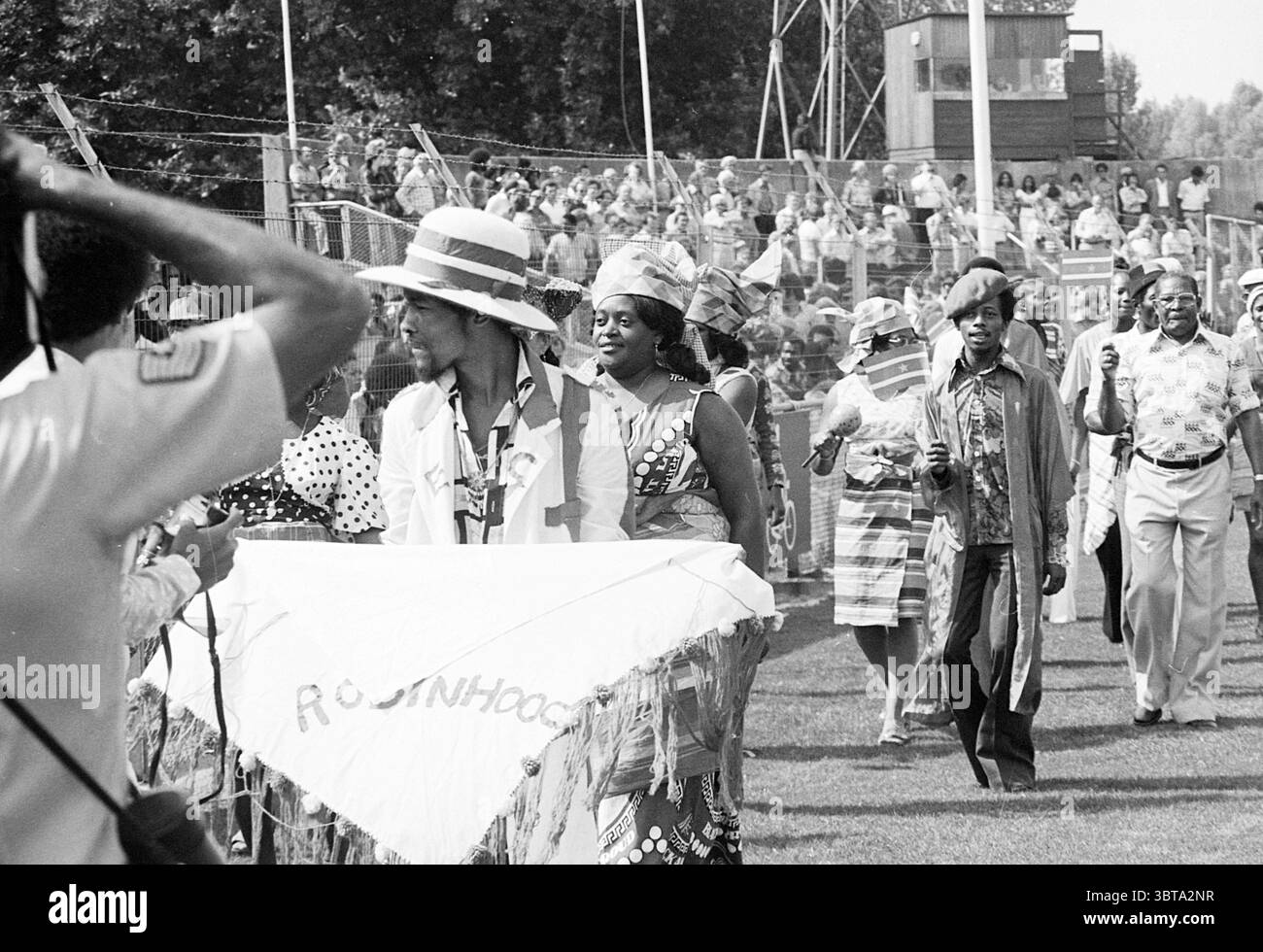 Haarlem - Robin Hood Football Haarlem, Whizgle News, Dutch Desk, Paesi Bassi, 1950 - 2000 il 15-08-1976. Questi sono gli elementi dell'immagine. La scena cattura una vivace ed energica processione, probabilmente durante un evento culturale o un festival. In primo piano, un gruppo di individui è prominente, ognuno decorato con abiti colorati che riflettono un ricco patrimonio culturale. Un personaggio si distingue con un grande cappello di paglia, mentre un altro indossa un outfit dal design complesso realizzato in tessuto a motivi geometrici. L'atmosfera è animata da un senso di celebrazione, poiché i partecipanti sembrano essere impegnati in vita Foto Stock
