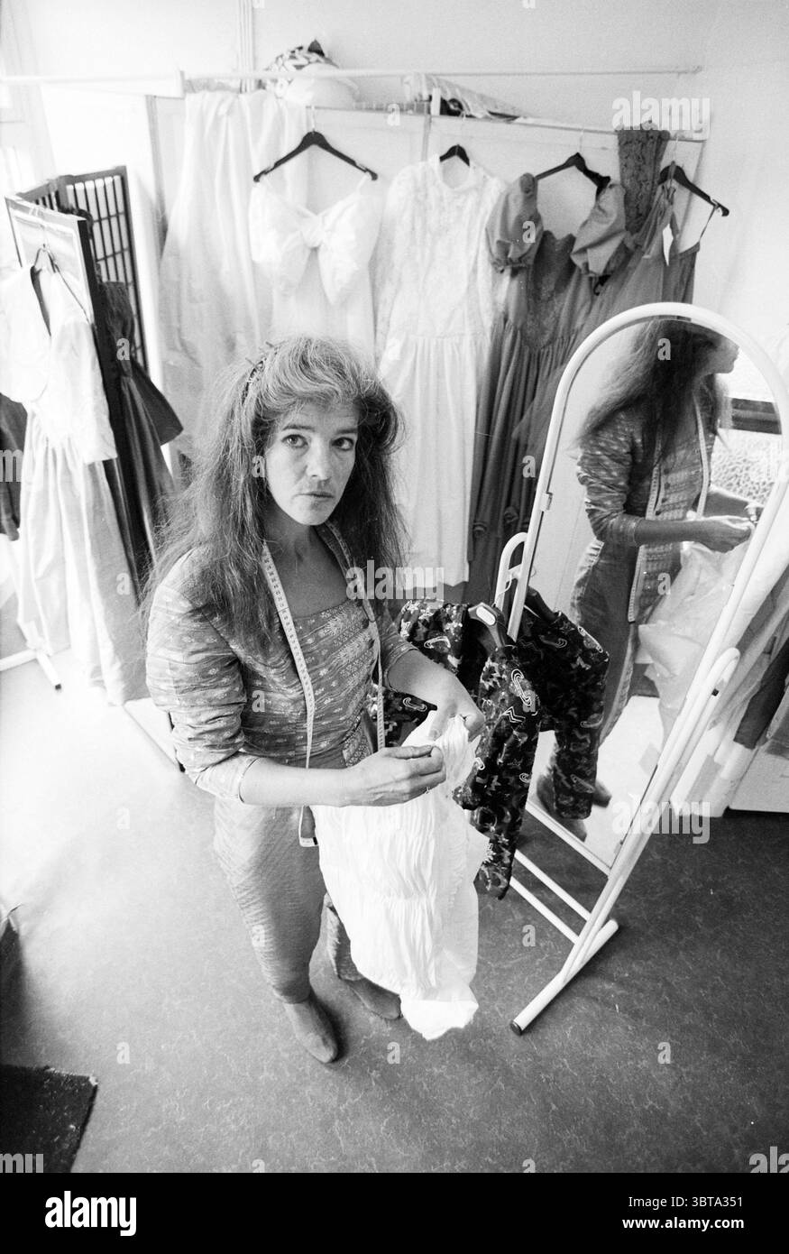 Jopie Schendel sarta, Whizgle News, Dutch Desk, Paesi Bassi, 1950 - 2000 il 11-06-1992. L'immagine mostra questi argomenti. La scena è ambientata in uno studio di abbigliamento pieno di indumenti e tessuti. Una persona sta in primo piano, guardando direttamente lo spettatore. Questa persona ha capelli lunghi e voluminosi con un leggero movimento ondulato, creando un contrasto sorprendente con le linee morbide del tessuto che sta tenendo. Indossati con top aderenti e pantaloni testurizzati, trasudano un look creativo ma al contempo mirato. Intorno a loro vi sono vari indumenti appesi su scaffali, tra cui abiti eleganti in soffice, Foto Stock