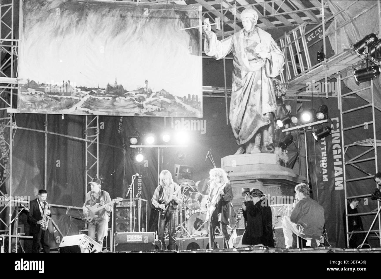 Gruppo pop Alexander su gr. Market Music, Whizgle News, Dutch Desk, Paesi Bassi, 1950 - 2000 il 13-06-1987. Questi argomenti sono mostrati nell'immagine. La scena è ambientata in un vivace luogo all'aperto, possibilmente uno spazio per festival o concerti. Sullo sfondo si infila una statua di spicco, raffigurante una figura storica con un'espressione poderosa ma nobile, che suggerisce un legame con la cultura o il patrimonio culturale. Questa statua si erge in cima a un piedistallo, circondato da una struttura a impalcatura che sorregge un ampio sfondo, caratterizzato da una rappresentazione artistica di paesaggi e forse un cielo spettacolare. In primo piano, una banda pe Foto Stock
