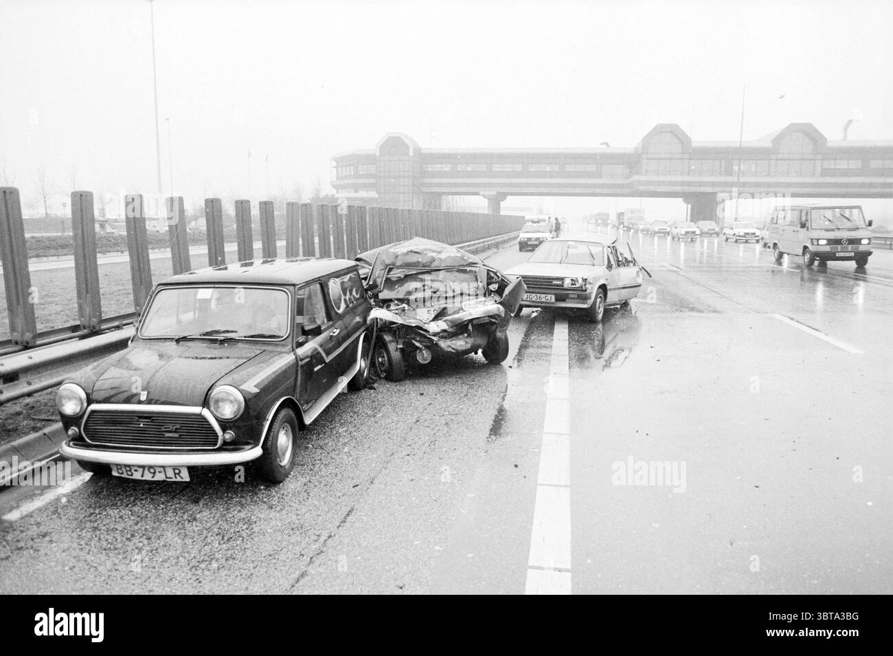 Incidente d'auto dovuto a condizioni scivolose, Whizgle News, Dutch Desk, Paesi Bassi, 1950 - 2000 il 02-1987. Questi sono gli argomenti nell'immagine. La scena cattura un'atmosfera cupa e cupa, caratterizzata da cieli ricoperti che creano uno sfondo grigio e silenzioso. L'ambiente sembra essere un'autostrada trafficata, con un lungo tratto di asfalto che riflette la pioggia leggera, che conferisce una lucentezza lucente alla superficie. In primo piano, due auto sono coinvolte in una collisione. Il primo veicolo a sinistra è un modello classico e compatto, identificabile dalla sua forma arrotondata e dalle caratteristiche retrò. Mostra un Foto Stock