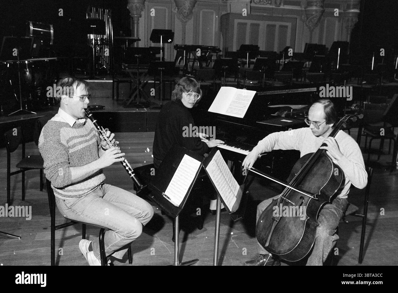 3 membri dell'orchestra NPO North Holland Philharmonic Orchestra, Whizgle News, Dutch Desk, Paesi Bassi, 1950 - 2000 il 12-04-1989. L'immagine mostra questi argomenti. La scena cattura tre musicisti impegnati nella pratica all'interno di una spaziosa sala. L'ambientazione è caratterizzata da un grande palcoscenico con file di sedie vuote disposte in previsione di un pubblico, suggerendo una disposizione del concerto. In primo piano, un uomo si siede su uno sgabello, giocando un oboe. Indossa occhiali e un maglione accogliente e dai colori chiari, creando un'atmosfera rilassata. Accanto a lui, una donna è posizionata ad un pianoforte, le sue dita sono addossate sopra il Foto Stock