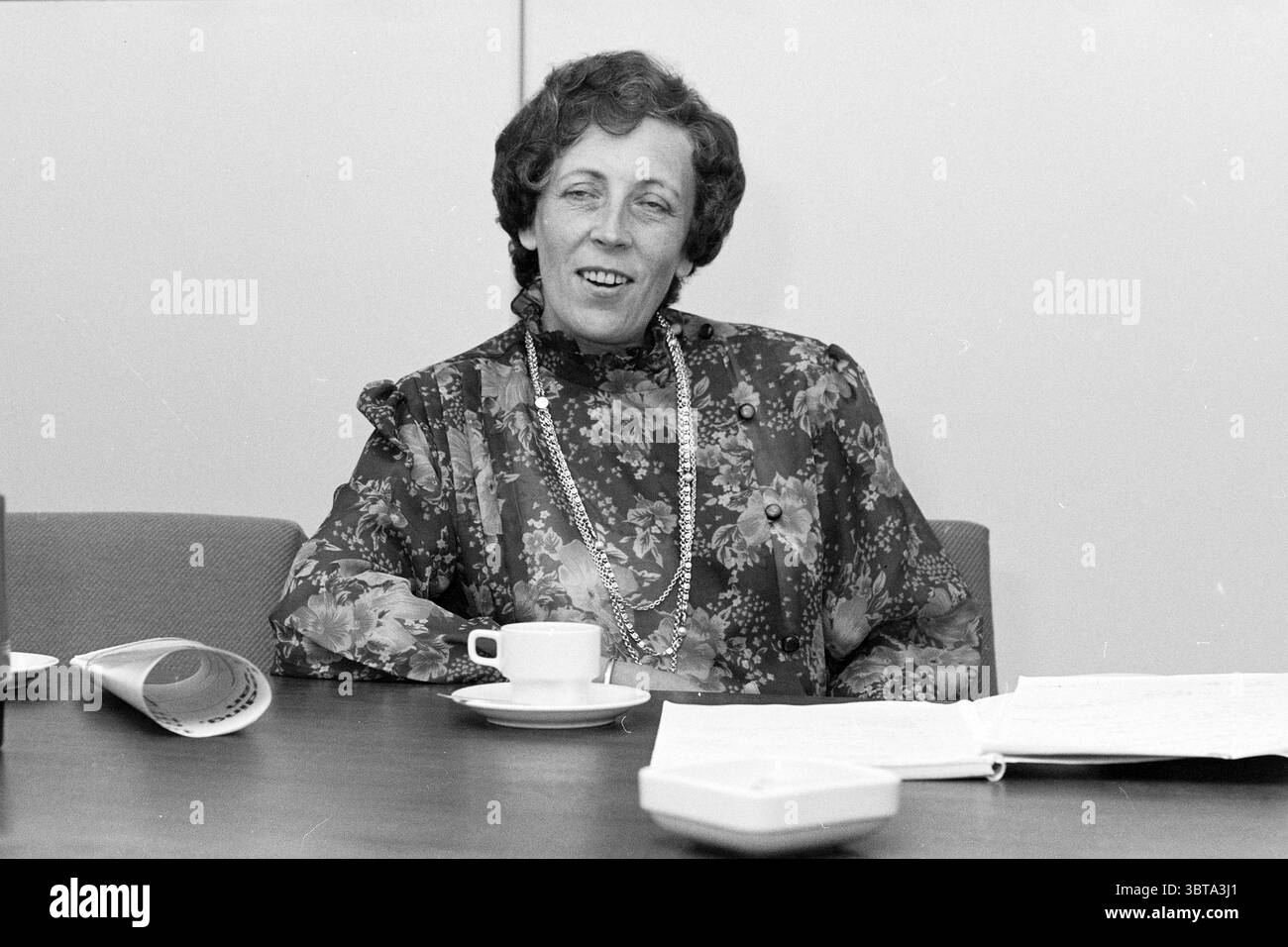 Sig.ra Appeldoorn Dietician Prov. Ospedale cibo e mangiare case Medicina metodo di guarigione, Whizgle News, Dutch Desk, Paesi Bassi, 1950 - 2000 il 06-10-1987. Questi argomenti sono mostrati nell'immagine. La scena vede una donna seduta a un tavolo, impegnata in una conversazione. Ha capelli ricci di media lunghezza e indossa una camicetta con un audace motivo floreale in vari colori, prevalentemente tonalità scure con sfumature più luminose. Intorno al collo c'è una lunga collana che aggiunge un tocco di eleganza al suo outfit. Il tavolo è in legno e presenta una superficie liscia, che riflette un tono caldo e ricco. Di fronte a lui Foto Stock