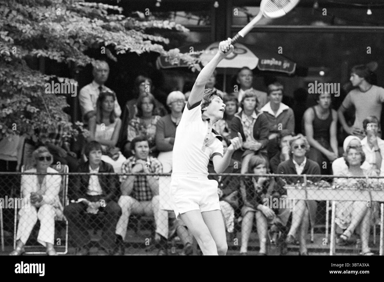 Associazione di tennis TVB Barendrecht., Whizgle News, Dutch Desk, Paesi Bassi, 1950 - 2000. Questi argomenti vengono visualizzati nell'immagine. In un ambiente all'aperto vivace, un giovane atleta viene catturato a metà corsa, pronto a servire una palla da tennis. Indossato con un classico abito da tennis bianco, che accentua le linee pulite della sua forma, si erge su un campo ben tenuto. La sua presa sulla racchetta suggerisce concentrazione e determinazione, e la posizione elevata del braccio indica un potente movimento che sta per svolgersi. Intorno al campo, una folla diversificata è seduta, mostrando una serie di reazioni, dall'eccitazione all'anticipazione Foto Stock