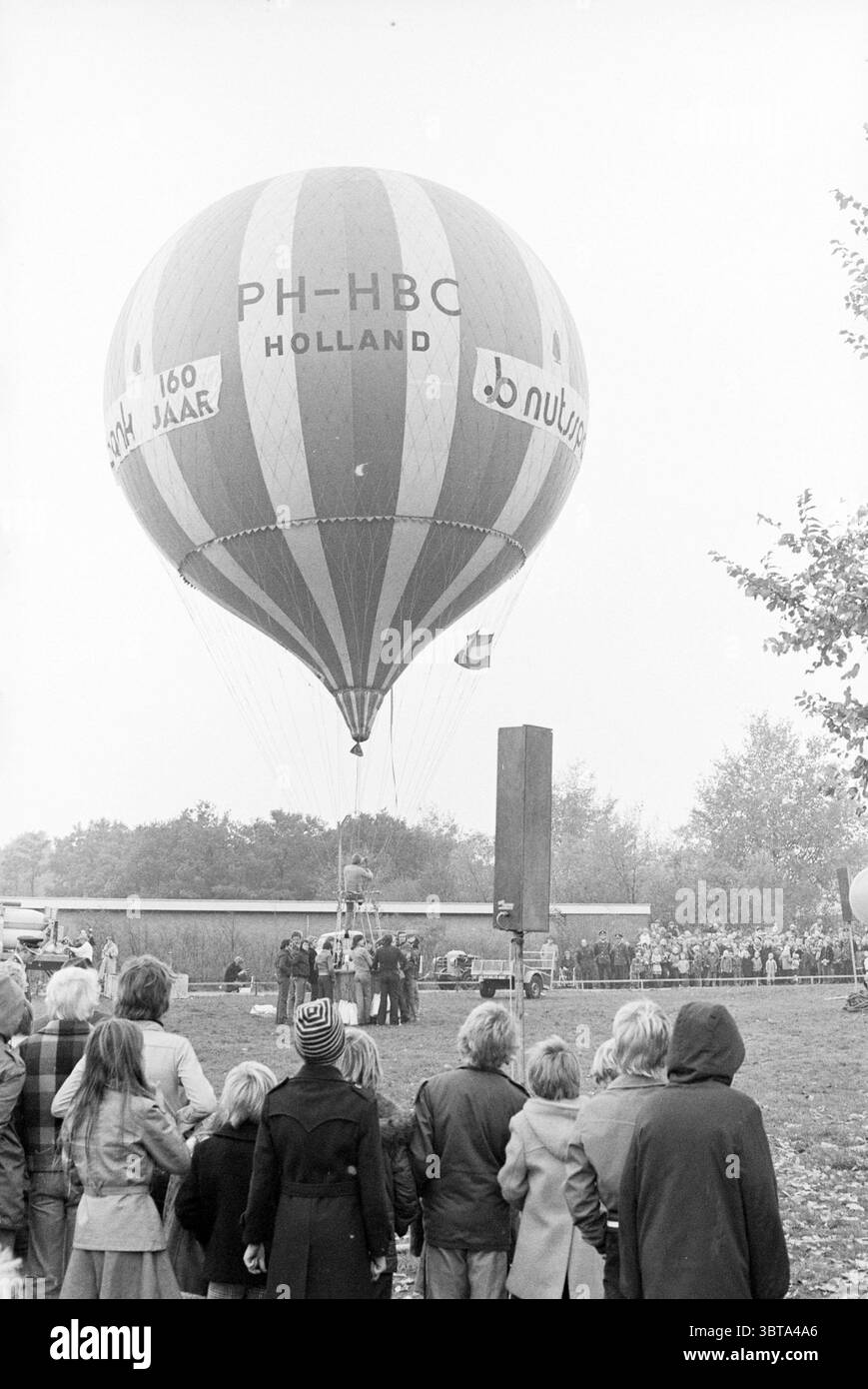 Mongolfiera Utility Savings Bank 160 anni, Whizgle News, Dutch Desk, Paesi Bassi, 1950 - 2000. Questi sono gli argomenti nell'immagine. In un ambiente tranquillo all'aperto, una vivace mongolfiera domina la scena, la sua forma rotonda adornata da audaci strisce rosse e bianche e contrassegnata con il nome "HOLLAND". Il pallone è parzialmente gonfiato vicino al suolo, emanando un senso di attesa mentre si prepara per l'ascesa. Riuniti intorno alla mongolfiera, un gruppo eterogeneo di persone è in attesa. Mostrano una varietà di stili di abbigliamento, indicativi di un'epoca precedente, con alcuni individui wearin Foto Stock