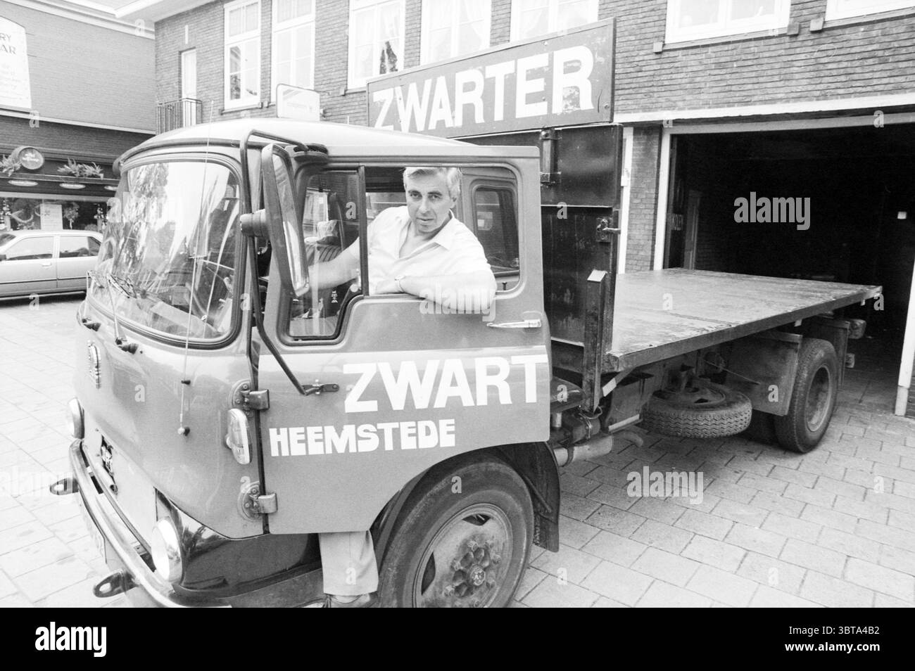 Camion del commercio di carbone Zwarter di Heemstede. Heemstede Raadhuisstraat Paesi Bassi, Whizgle News, Dutch Desk, Paesi Bassi, 1950 - 2000 il 10-07-1987. Questi argomenti vengono visualizzati nell'immagine. Nella scena, un camion d'epoca occupa il primo piano, la sua vernice rosso brillante contrasta con i toni tenui degli edifici in mattoni sullo sfondo. La cabina del camion è aperta, rivelando un uomo seduto sul sedile di guida, che si appoggia casualmente alla porta. Ha un comportamento rilassato, con un leggero sorriso, che suggerisce un'interazione amichevole con chiunque nelle vicinanze. Il carrello ha una superficie piana e la superficie in legno è Foto Stock
