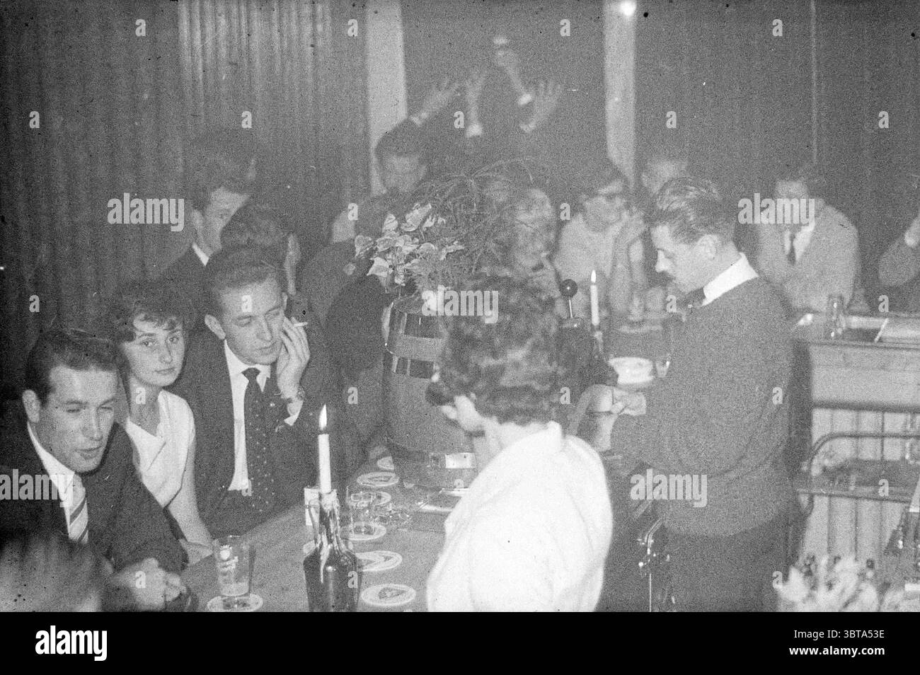 Barista e interno 'De Grot' Cafe, Whizgle News, Dutch Desk, Paesi Bassi, 1950 - 2000 il 18-08-1961. Questi sono gli argomenti nell'immagine. La scena cattura un ambiente bar vivace e poco illuminato pieno di persone impegnate in conversazione e socializzazione. In prima linea, un gruppo di individui occupa un lungo tavolo. L'ambiente suggerisce un'atmosfera vintage, con calde tonalità di marrone e sfumature più profonde che creano un'atmosfera intima ma vivace. Le persone a tavola esprimono varie espressioni: Alcune sorridono e animano, mentre altre appaiono contemplative, perdute nel pensiero. Una giovane donna con Foto Stock