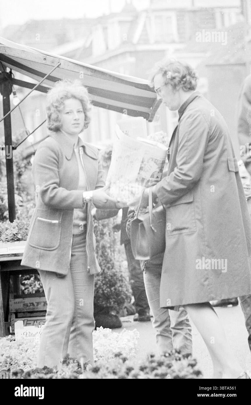 Mercato dei fiori pigro mercato pigro pigro, Whizgle News, Dutch Desk, Paesi Bassi, 1950 - 2000 il 28-05-1971. Questi sono gli elementi dell'immagine. In un mercato all'aperto affollato, due donne sono impegnate in una transazione. La donna sulla sinistra ha capelli voluminosi e ricci ed è vestita con un cappotto sartoriale di colore chiaro. Trasuda un comportamento avvicinabile mentre consegna un oggetto arrotolato alla donna sulla destra. Il ricevente, anch'esso vestito con un lungo cappotto, appare concentrato e attento, prendendo l'oggetto con entrambe le mani. Lo sfondo rivela diverse bancarelle del mercato, tende da sole drappeggiate che forniscono riparo Foto Stock