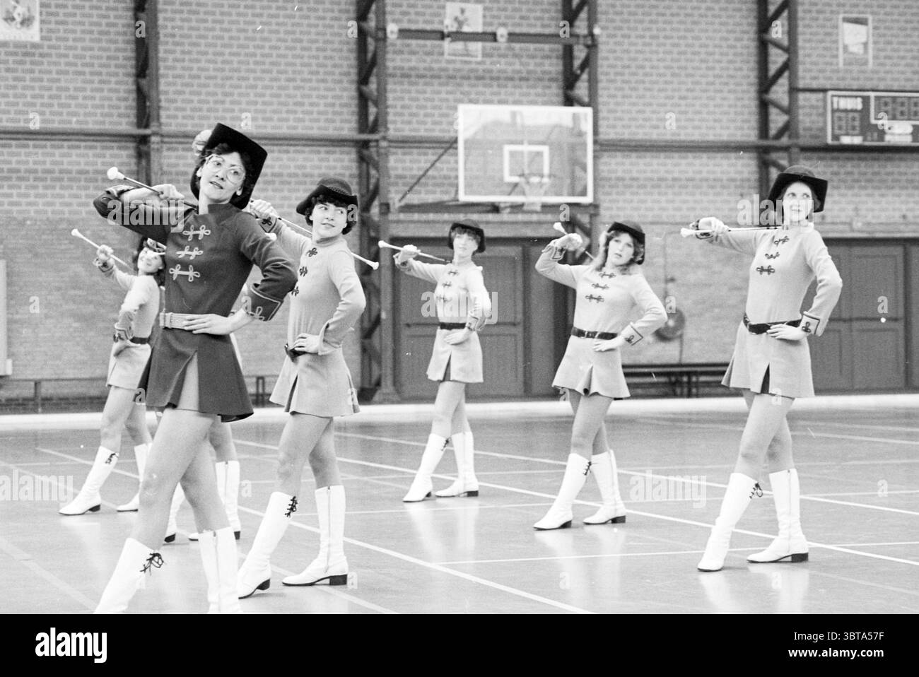 Majorette competition Kennemer Sports hall Majorettes, Whizgle News, Dutch Desk, Paesi Bassi, 1950 - 2000 il 25-05-1980. Questi argomenti vengono visualizzati nell'immagine. In una spaziosa palestra, sei artisti sono disposti in fila, mostrando una danza o una routine sincronizzata. L'ambiente presenta alti soffitti, pavimenti in legno e pareti in mattoni che gli danno un'atmosfera utilitaria, tipica di una sala sportiva. I ballerini sono vestiti con un abbigliamento coordinato che combina una vibrante tonalità di rosso con accenti contrastanti, creando un impatto visivo sorprendente. Ogni outfit presenta un corpetto aderente, svasatura in vita, e. Foto Stock