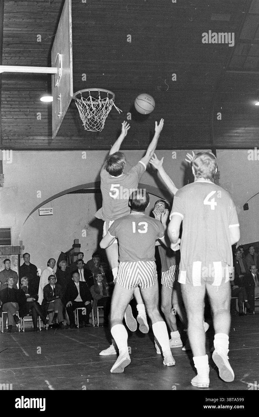 Partita di basket., Whizgle News, Dutch Desk, Paesi Bassi, 1950 - 2000. L'immagine mostra questi argomenti. In un ambiente interno vivace, si svolge una partita di basket caratterizzata da un'atmosfera dinamica. La palestra presenta un alto soffitto ad arco in legno che aggiunge calore alla scena. Il pavimento in legno lucido riflette le luci dall'alto, aumentando l'energia del momento. In primo piano, i giocatori che indossano maglie rosse con i numeri "5", "4" e "13" si impegnano in modo evidente in una partita tesa. L'atleta che indossa la maglia numero "5" salta in alto, raggiungendo il basket, il suo sculo Foto Stock
