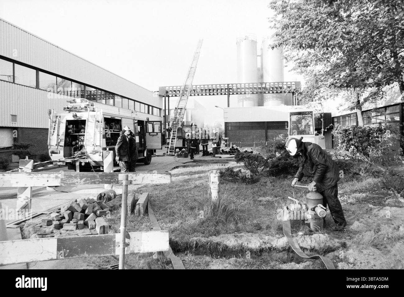 Esercitazione regionale dei vigili del fuoco MSD (compreso il collegamento dell'idrante del fuoco) Haarlem Paesi Bassi, Whizgle News, Dutch Desk, Paesi Bassi, 1950 - 2000 il 23-05-1989. Questi argomenti vengono visualizzati nell'immagine. La scena presenta un ambiente industriale affollato, composto principalmente da un grande edificio con un esterno in metallo ondulato verniciato in colori chiari. Diverse strutture cilindriche alte si innalzano sullo sfondo, indicando un impianto di produzione o di lavorazione. In primo piano, viene utilizzato un idrante del fuoco, con un vigile del fuoco vestito con equipaggiamento scuro che si inginocchia accanto ad esso. Il pugile del fuoco Foto Stock