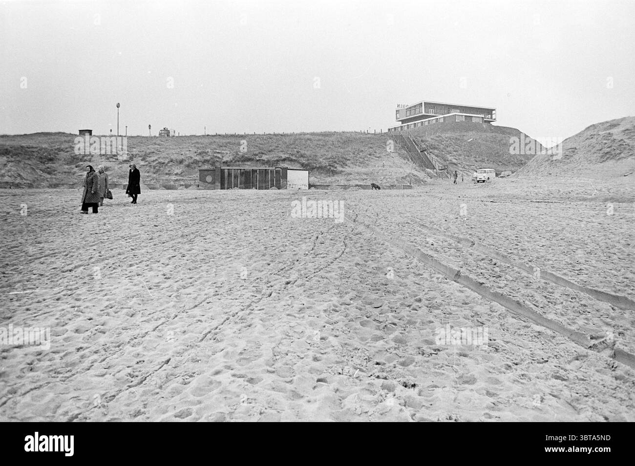 Ingresso alla spiaggia di Wijk aan Zee. Spiaggia e spiagge, Whizgle News, Dutch Desk, Paesi Bassi, 1950 - 2000 il 05-03-1975. Questi sono gli argomenti nell'immagine. La scena cattura un'ampia spiaggia sabbiosa sotto un cielo soffice e coperto, donando all'atmosfera una qualità cupa ma serena. La sabbia è contrassegnata da una serie di passi e tracce di pneumatici che conducono verso l'orizzonte lontano. In primo piano, due figure vestite di abiti scuri passeggiano per la spiaggia, sottolineando la loro presenza contro la sabbia dai toni chiari. Una figura cammina lungo il lato sinistro, mentre il loro cappotto si libra leggermente nella brezza Foto Stock