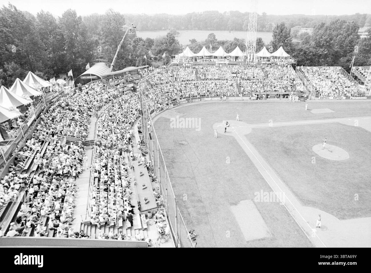Panoramica dello stadio Haarlem Baseball Week dall'elicottero Haarlem Paesi Bassi, Whizgle News, Dutch Desk, Paesi Bassi, 1950 - 2000 il 22-07-1990. Questi argomenti vengono visualizzati nell'immagine. La scena cattura un vivace stadio di baseball pieno di spettatori. Gli stand sono densamente popolati e presentano una folla diversificata di abbigliamento estivo. Cappelli da sole, camicie casual e pantaloncini corti dominano l'abbigliamento, suggerendo una giornata calda. Molti partecipanti sono seduti, mentre alcuni sono in piedi, aggiungendo un'atmosfera vivace. Il colore gioca un ruolo discreto in questo ambiente monocromatico, in cui le sfumature di grigio creano un fastidio Foto Stock