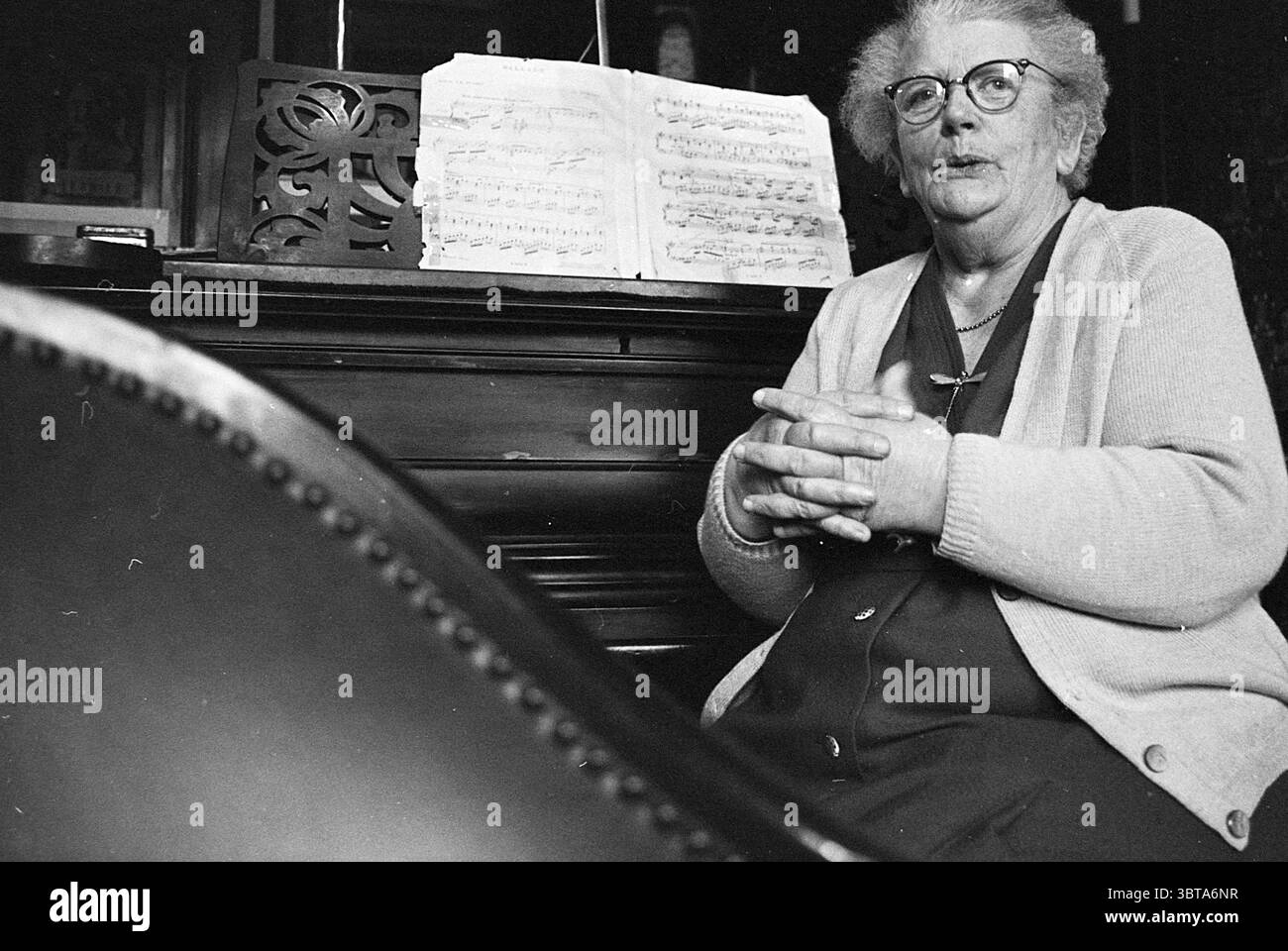 Pianista E. van Ede Music, Whizgle News, Dutch Desk, Paesi Bassi, 1950 - 2000 il 21-01-1971. Questi argomenti vengono visualizzati nell'immagine. La scena vede una donna anziana seduta vicino a un pianoforte, creando un'atmosfera calda e nostalgica. Ha i capelli grigi rifiniti con cura e indossa occhiali rotondi che accentuano la sua espressione gentile. Vestita con un cardigan chiaro sopra un abito scuro, le sue mani sono legate tra loro in grembo, trasmettendo un senso di calma e di portamento. In sottofondo, gli spartiti musicali sono esposti su un banco musicale, suggerendo un interesse o una pratica musicale. Le lenzuola, leggermente stropicciate, suggeriscono previ Foto Stock