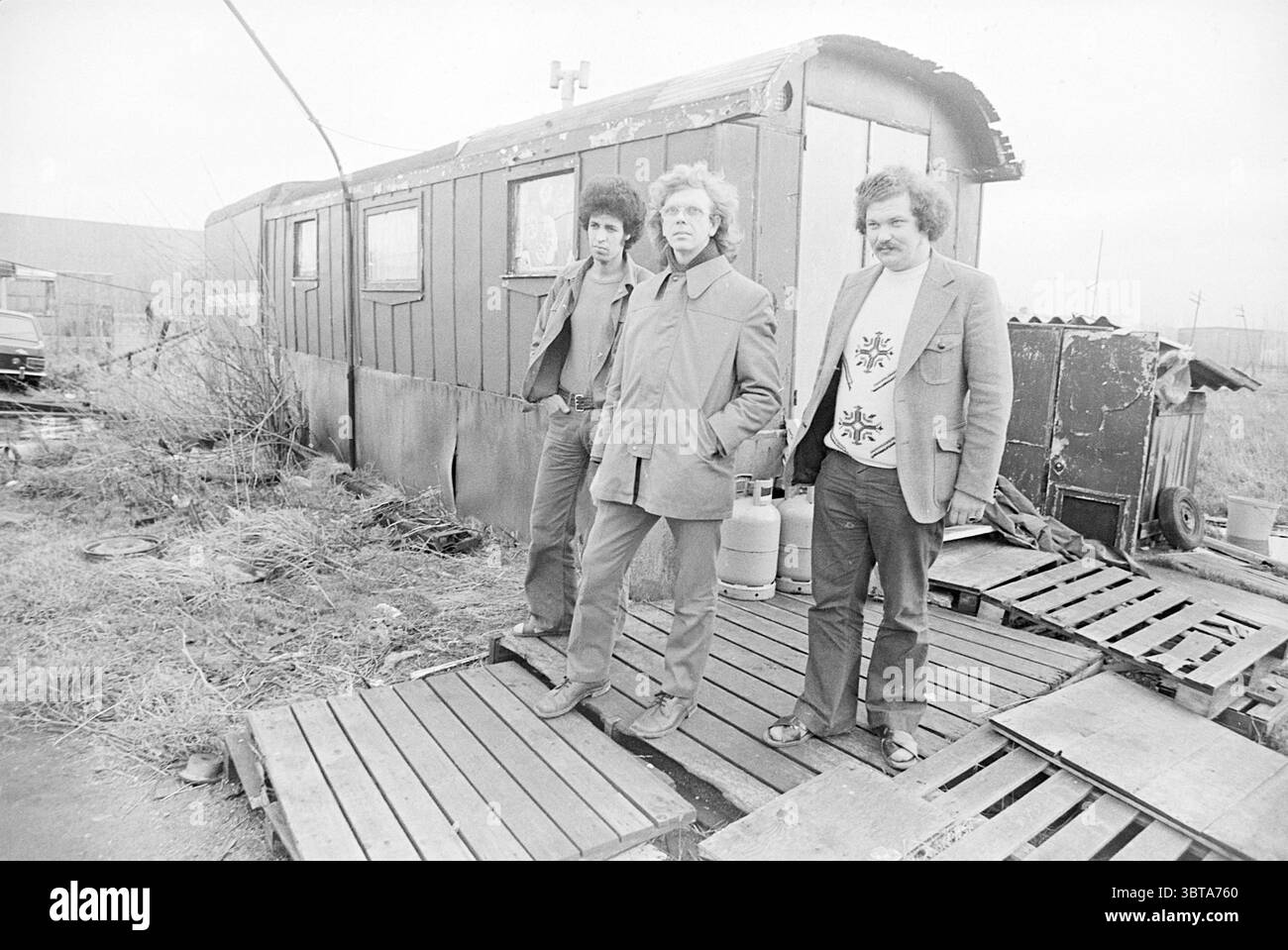 Hotel Maroccan Dam Factory Hoofddorp Guest Workers, Whizgle News, Dutch Desk, Paesi Bassi, 1950 - 2000 il 07-01-1976. Questi sono gli elementi dell'immagine. La scena cattura tre uomini in piedi su pallet di legno all'esterno di un rimorchio intempestivo. Il rimorchio, verniciato in toni tenui di grigio e marrone, mostra segni di età con vernice sbucciante e un tetto inclinato, che si fonde nel cielo coperto che proietta una luce diffusa sull'area. Gli uomini mostrano stili distinti: Uno ha capelli ricci scuri ed è vestito in modo informale, indossando una giacca e un jeans. Il secondo uomo sta al centro, con un po' Foto Stock