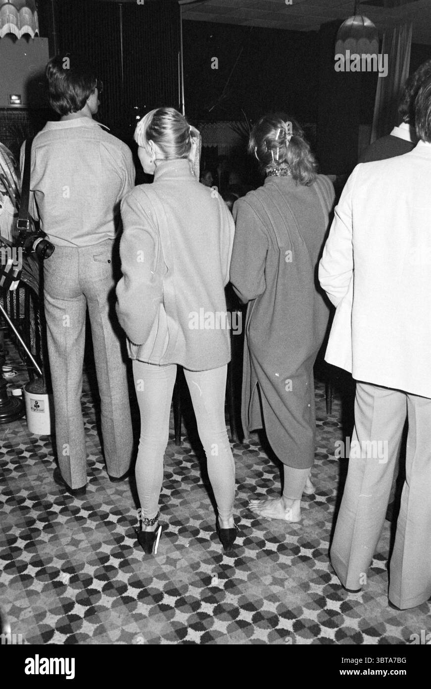 Exam Evening Models Agency Wintertuin Exam Models, Whizgle News, Dutch Desk, Paesi Bassi, 1950 - 2000 il 08-12-1979. Questi argomenti sono mostrati nell'immagine. La scena cattura un momento in un ambiente interno vivace, probabilmente un evento sociale o di riunione. In primo piano, tre figure si avvicinano, le spalle si rivolgono allo spettatore, suggerendo un'esperienza condivisa o focalizzarsi su un punto focale invisibile. La prima figura a sinistra è vestita con un abito grigio chiaro, il tessuto morbido e sartoriale conferisce un aspetto raffinato. I capelli dell'individuo sono ben modellati, contribuendo alla Foto Stock