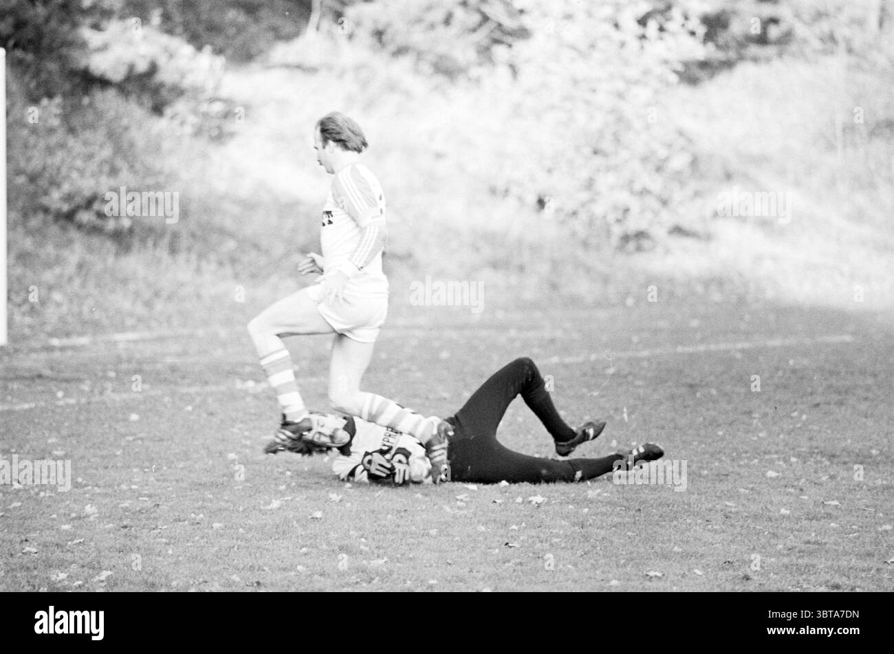 SVB - Bloemendaal Football, Whizgle News, Dutch Desk, Paesi Bassi, 1950 - 2000 il 21-11-1982. L'immagine mostra questi argomenti. In una dinamica scena all'aperto su un campo erboso, l'azione cattura un momento in una partita di calcio. In primo piano sono presenti due giocatori impegnati in un'interazione fisica. Un giocatore, vestito con un'uniforme di colore chiaro con strisce verticali, è raffigurato a mezza riga, sollevato dal suolo, suggerendo movimento e agilità. Il giocatore appare concentrato, con determinazione evidente nella sua postura. Sotto di loro si trova un altro giocatore vestito con abiti scuri, sparsi per terra, sugg Foto Stock