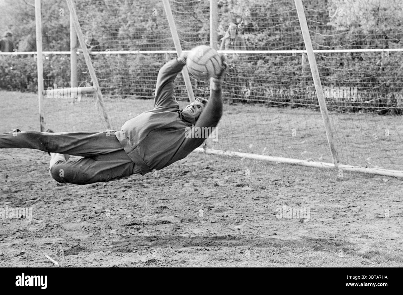 Jaap Bloem di Telstar Football People and Trainer, Whizgle News, Dutch Desk, Paesi Bassi, 1950 - 2000 on 25-04-1974. Questi argomenti sono mostrati nell'immagine. La scena cattura un momento dinamico su un campo sportivo, in cui un giocatore si sta tuffando per prendere una palla da calcio. Il capo è vestito con una camicia a maniche lunghe e pantaloni aderenti, per un comfort e un'agilità eccezionali. Il loro corpo è arcuato graziosamente a mezz'aria, mostrando una miscela di determinazione e atletismo. Lo sfondo presenta un semplice parco giochi, con un'altalena e un'area con rete, che indica uno spazio ricreativo. Intorno alla f Foto Stock
