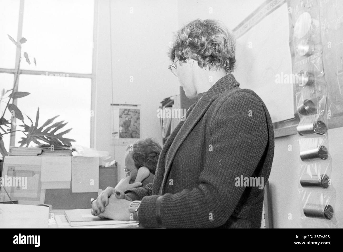 Donna in ufficio con lavagna scritta, Whizgle News, Dutch Desk, Paesi Bassi, 1950 - 2000 il 13-10-1982. L'immagine contiene questi argomenti. La scena presenta due persone in un ambiente interno che trasuda un'atmosfera tranquilla e accademica. Una persona, posizionata in primo piano, ha una postura premurosa, guardando leggermente di lato. Indossano un cappotto scuro e testurizzato che aggiunge un tocco di raffinatezza, completato da occhiali rotondi che impreziosiscono la loro espressione attenta. Sullo sfondo, un secondo individuo sembra essere impegnato in attività, parzialmente oscurato dalla vista dai primi pers Foto Stock