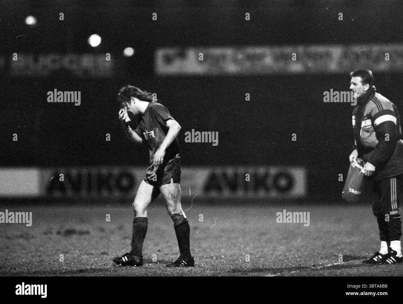 Partita di calcio Haarlem, Whizgle News, Dutch Desk, Paesi Bassi, 1950 - 2000 nel 1988. Questi argomenti vengono visualizzati nell'immagine. La scena si sviluppa su un campo da calcio poco illuminato, avvolto da un'atmosfera cupa. L'atmosfera sembra pesante, probabilmente a causa di una giocata recente o di uno sfortunato incidente in campo. In primo piano, un giocatore si allontana dall'azione, si abbassa la testa e si copre la mano, suggerendo una dedizione o una delusione. La sua maglia è scura, a indicare uno stile di calcio classico, mentre i suoi pantaloncini appaiono leggermente fangosi, a indicare una recente lotta con gli elementi del gioco. Il pla Foto Stock