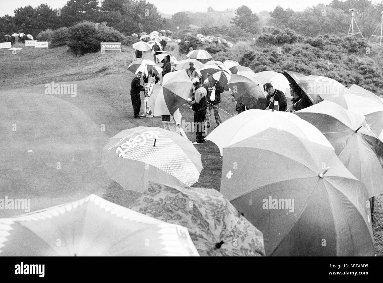 KLM apre varie registrazioni Golf Golf Golf 29-07-1989, Whizgle News, Dutch Desk, Paesi Bassi, 1950 - 2000 il 30-07-1989. L'immagine mostra questi argomenti. La scena è ambientata su un campo da golf sotto un cielo grigio e coperto, suggerendo una giornata di pioggia. Numerosi individui sono riuniti sulla lussureggiante erba verde del campo, tutti con grandi ombrelli, che creano un baldacchino colorato sopra di loro. Gli ombrelli hanno un design vario, alcuni presentano motivi audaci e tonalità luminose, mentre altri sono più silenziosi e semplici. In primo piano, un gruppo di persone è stretto insieme, apparendo impegnato in co Foto Stock