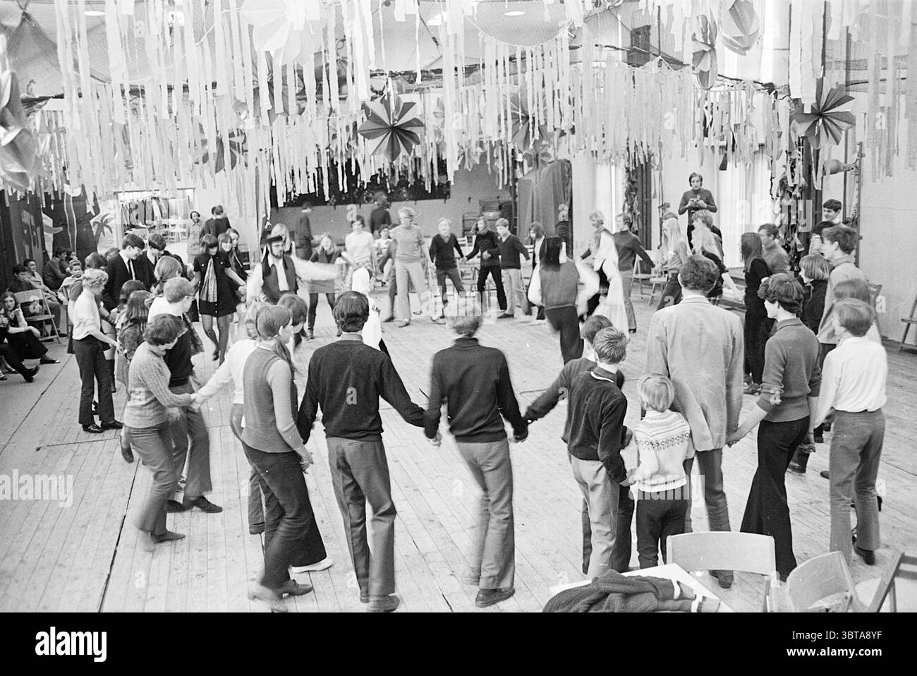 Scuola di grammatica di danza popolare scuole di danza popolare, Whizgle News, Dutch Desk, Paesi Bassi, 1950 - 2000 il 20-03-1970. Questi sono gli argomenti nell'immagine. In una vivace scena interna, un gruppo di persone si riunisce in una spaziosa sala adornata da rastrelli colorati e decorazioni che pendono dal soffitto, creando un'atmosfera festosa. I colori vivaci dei rastrelli contrastano con le tonalità di legno naturale del pavimento, che ha un aspetto caldo e rustico. Al centro, i partecipanti formano un grande cerchio, tenendosi per mano e impegnandosi in una danza animata. Gli individui espongono una vasta gamma di abbigliamento Foto Stock