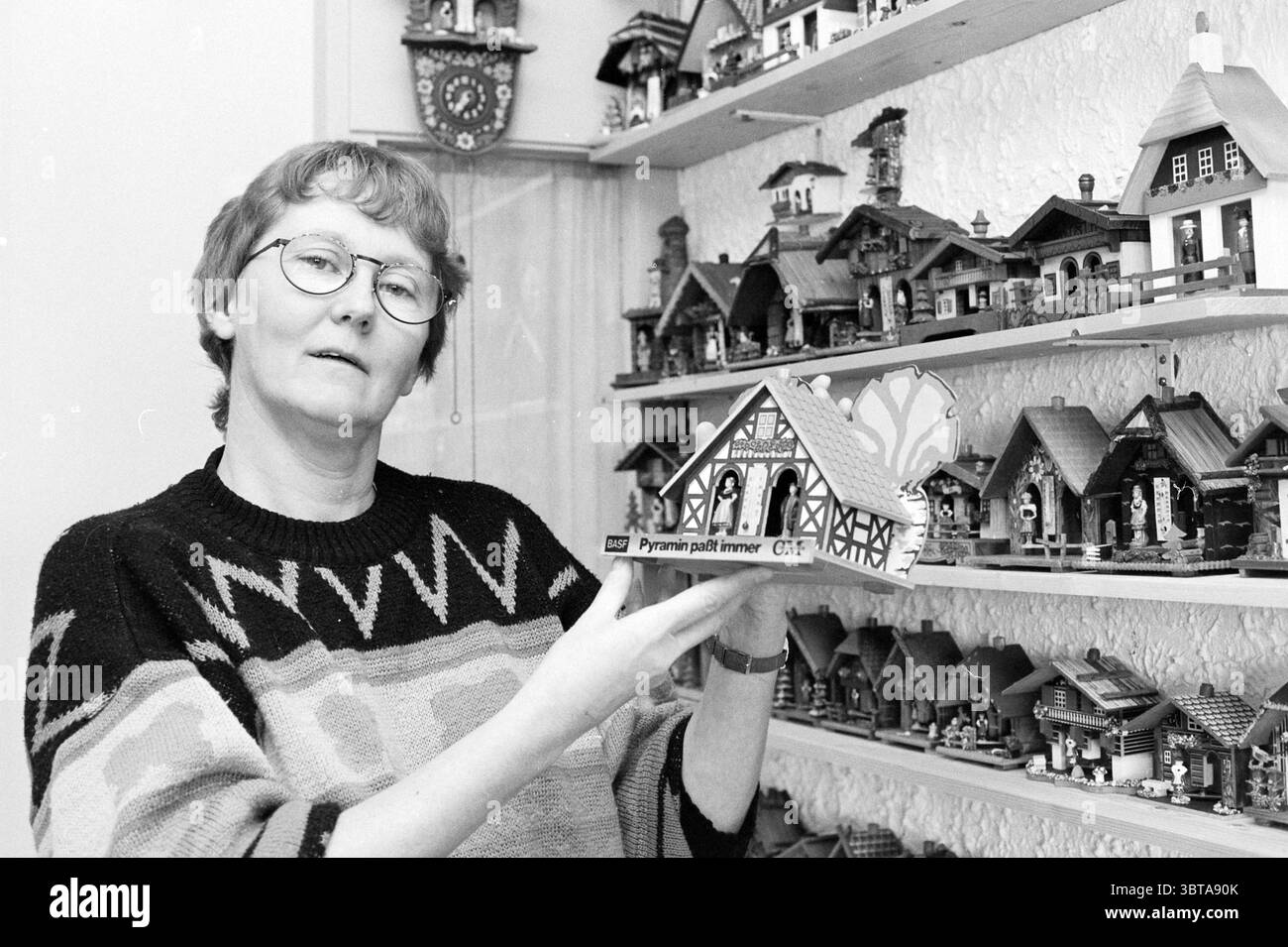 Mrs. Wittekoek Weather Houses, Whizgle News, Dutch Desk, Paesi Bassi, 1950 - 2000 il 13-12-1993. Questi sono gli argomenti nell'immagine. In un ambiente interno accogliente, una donna si erge in primo piano, tenendo in mano un modello decorativo di casa in legno. Indossa occhiali rotondi e un pullover elegante caratterizzato da un audace motivo geometrico, prevalentemente in tonalità di grigio con colori contrastanti. L'atmosfera è calda e invitante, suggerendo una passione per l'artigianato o il collezionismo. Dietro di lei, una serie di scaffali sono pieni di una serie di case in miniatura dal design complesso, che mostrano colori vivaci A. Foto Stock