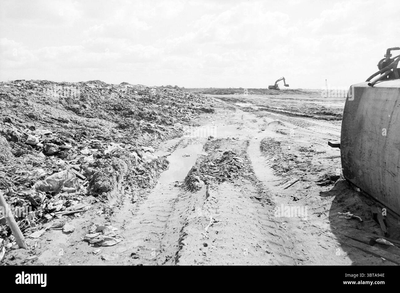 Garbage dump Aagtendijk Beverwijk Garbage Beverwijk Paesi Bassi, Whizgle News, Dutch Desk, Paesi Bassi, 1950 - 2000 il 30-06-1989. Questi argomenti vengono visualizzati nell'immagine. La scena raffigura un paesaggio desolato dominato da una vasta distesa di terreno irregolare. Il terreno è in gran parte composto da sporcizia e detriti, con pile sparse di rifiuti che includono un mix di rifiuti organici e vari materiali scartati. La superficie è caratterizzata da profondi binari di pneumatici che si snodano attraverso il sito, suggerendo recenti attività di macchinari pesanti. Sullo sfondo, un escavatore è grande, il suo potente braccio Foto Stock