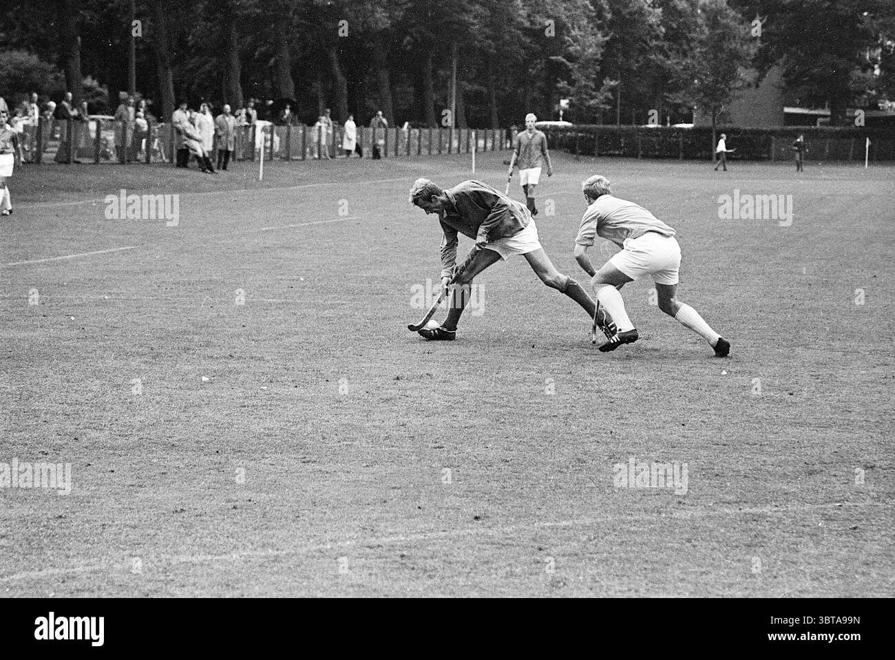 BMHC - Kampong Hockey, Whizgle News, Dutch Desk, Paesi Bassi, 1950 - 2000 il 03-09-1967. L'immagine include questi argomenti. La scena cattura un momento intenso su un lussureggiante campo verde definito da una superficie erbosa ben curata. Due atleti sono i punti focali, impegnati in un gioco dinamico. Un giocatore, rivestito di una maglia di colore scuro, si piega in basso mentre manovra abilmente un bastone da hockey, pronto a colpire la palla. La sua espressione focalizzata riflette determinazione e concentrazione. L'altro giocatore, che indossa una maglia di colore chiaro, è posizionato dietro di sé, mostrando una postura che suggerisce di esserlo Foto Stock