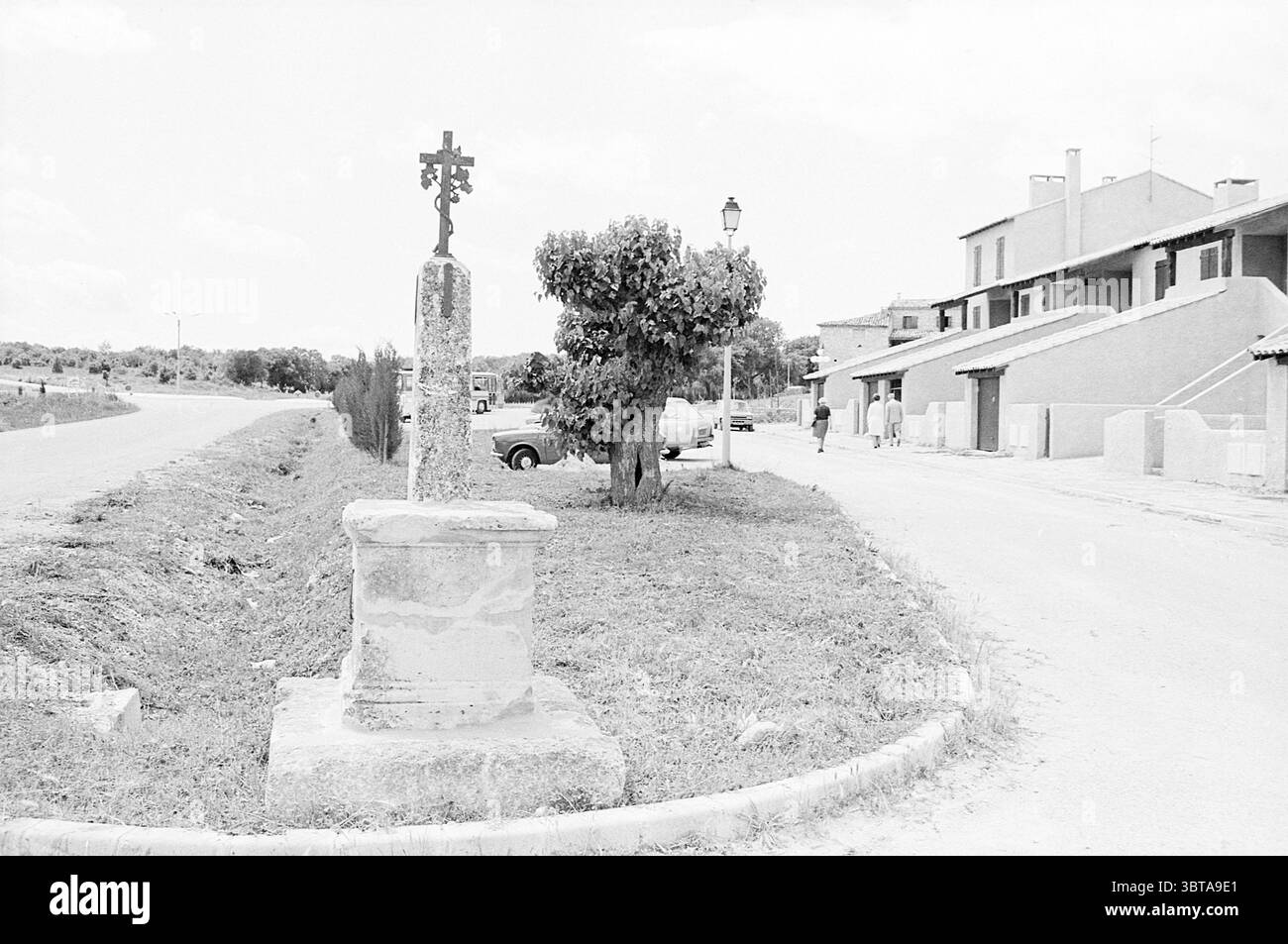 Relazione Mesjaunne Francia - pannelli solari per il calore relazione, Whizgle News, Dutch Desk, Paesi Bassi, 1950 - 2000 il 17-06-1975. Questi argomenti vengono visualizzati nell'immagine. La scena mostra un ambiente rurale lungo una strada tranquilla. Su un lato, una croce di pietra si erge in modo prominente su un piedistallo intempestivo, la sua consistenza ruvida e invecchiata, suggerendo che ha resistito agli elementi per molti anni. La croce stessa è adornata da intricate incisioni che aggiungono carattere al paesaggio. Dietro la croce, cresce un lussureggiante albero verde, le sue foglie gettano forme dappate sul terreno, che appaiono vibranti contro gli altri miti Foto Stock