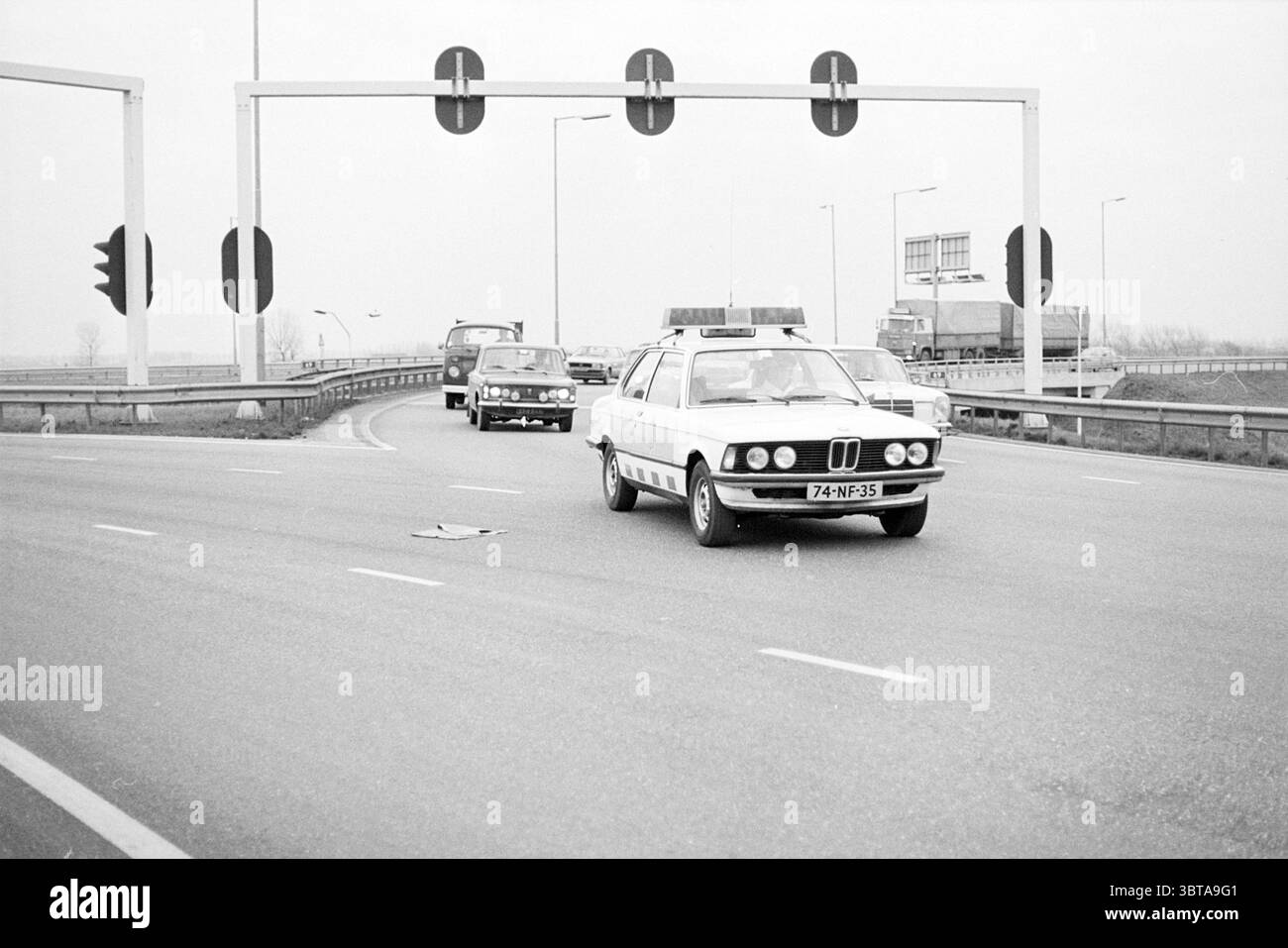 Polizia di sorveglianza Rijksweg IJm. Polizia, Whizgle News, Dutch Desk, Paesi Bassi, 1950 - 2000 il 02-12-1977. Questi sono gli argomenti nell'immagine. La scena cattura un ambiente di autostrada affollato sotto cieli ricoperti, dando un'atmosfera silenziosa e un po' cupa. Una classica auto della polizia bianca e blu, in primo piano, si distingue sullo sfondo in scala di grigi. E' dotata di una barra luminosa sul tetto, che aggiunge l'impressione di urgenza e autorevolezza. Sullo sfondo, un altro veicolo, di colore più scuro, corre dietro l'auto della polizia, mentre un camion più grande è di fronte Foto Stock