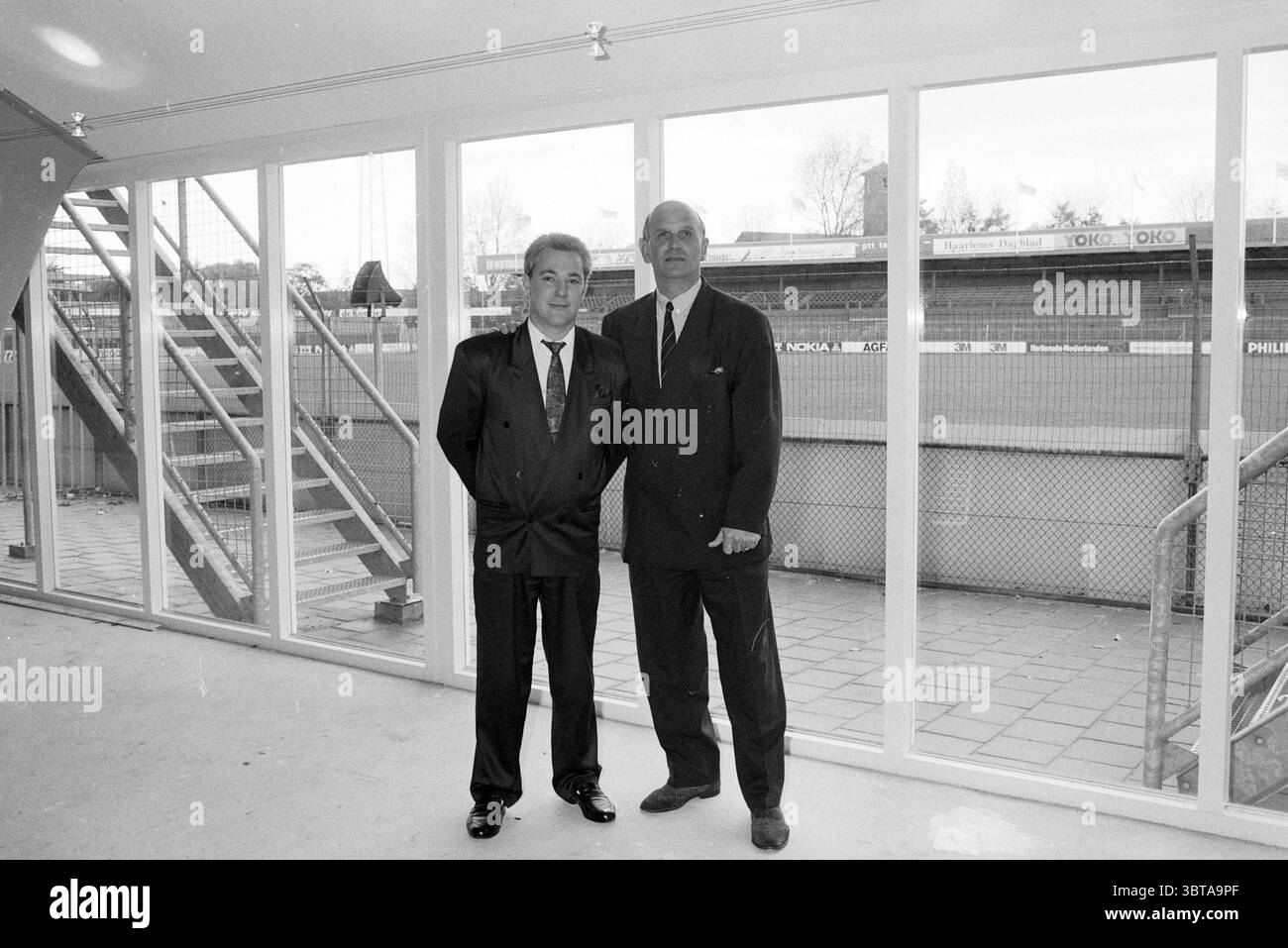 Barry Hughes e un altro uomo nello stadio di HFC Haarlem Haarlem Paesi Bassi, Whizgle News, Dutch Desk, Paesi Bassi, 1950 - 2000 il 10-1990. L'immagine mostra questi argomenti. La scena presenta due persone affiancate in un moderno spazio interno con grandi pannelli di vetro, che consentono alla luce naturale di inondare l'area. Lo sfondo rivela un campo sportivo, parzialmente oscurato dal vetro, che mostra erba verde e contorni di pali, che accennano a un luogo di atletica. Entrambe le persone sono vestite in abiti formali, con abiti che trasmettono professionalità. L'uomo a sinistra ha la zampa Foto Stock
