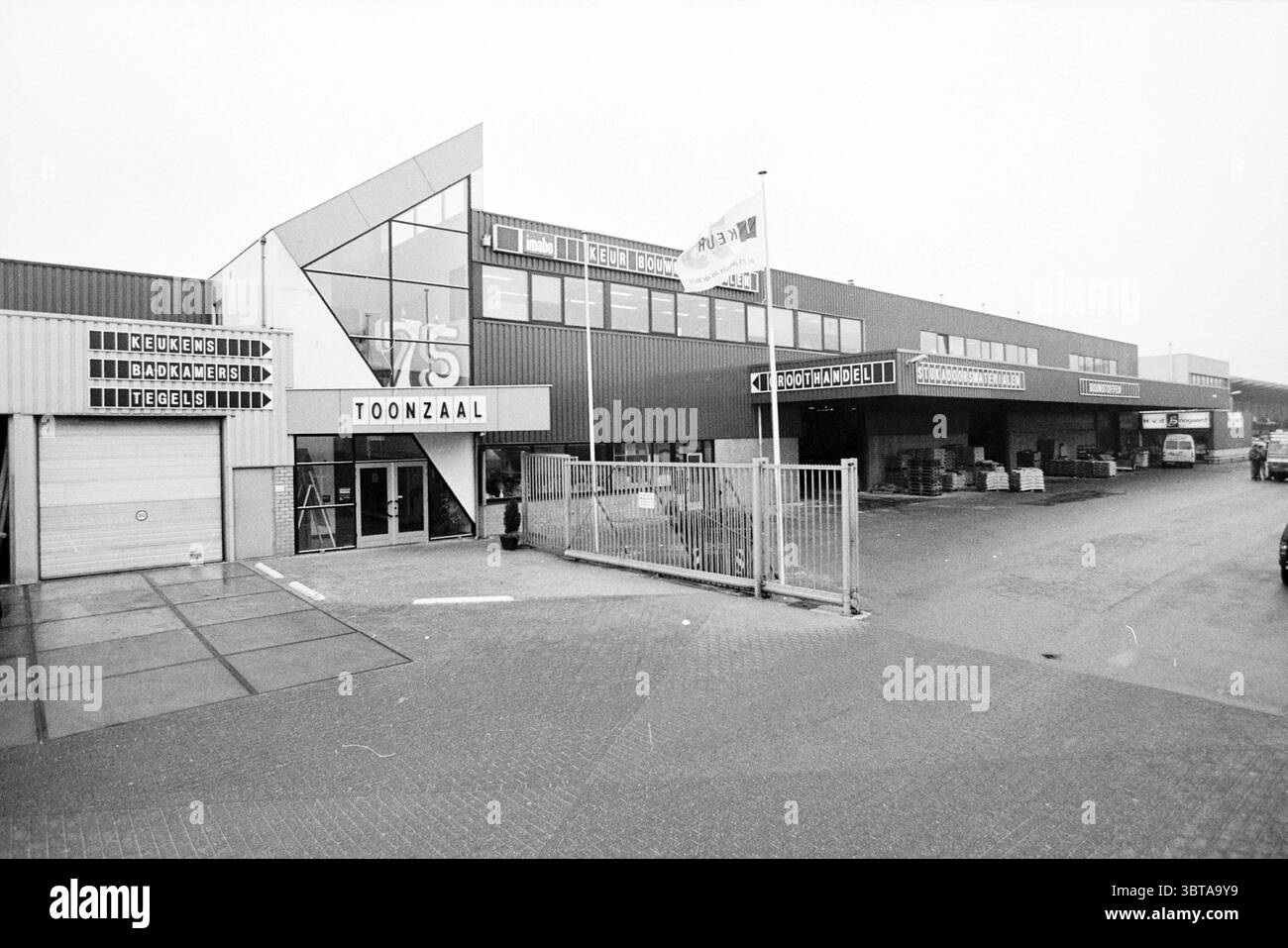 Exterior Inspection of Building Materials, Whizgle News, Dutch Desk, Paesi Bassi, 1950 - 2000 on 27-01-1998. Questi sono gli elementi dell'immagine. La scena raffigura un grande edificio industriale caratterizzato dal suo design architettonico moderno. L'esterno presenta una combinazione di materiali grigi e blu scuro che gli conferiscono un aspetto elegante e contemporaneo. Posizionato in modo prominente è un ingresso in vetro triangolare, che aggiunge un elemento geometrico impressionante alla struttura. Di fronte a questa entrata c'è una recinzione metallica che separa il passaggio pedonale dall'area parcheggio. La superficie del terreno è pavimentata Foto Stock