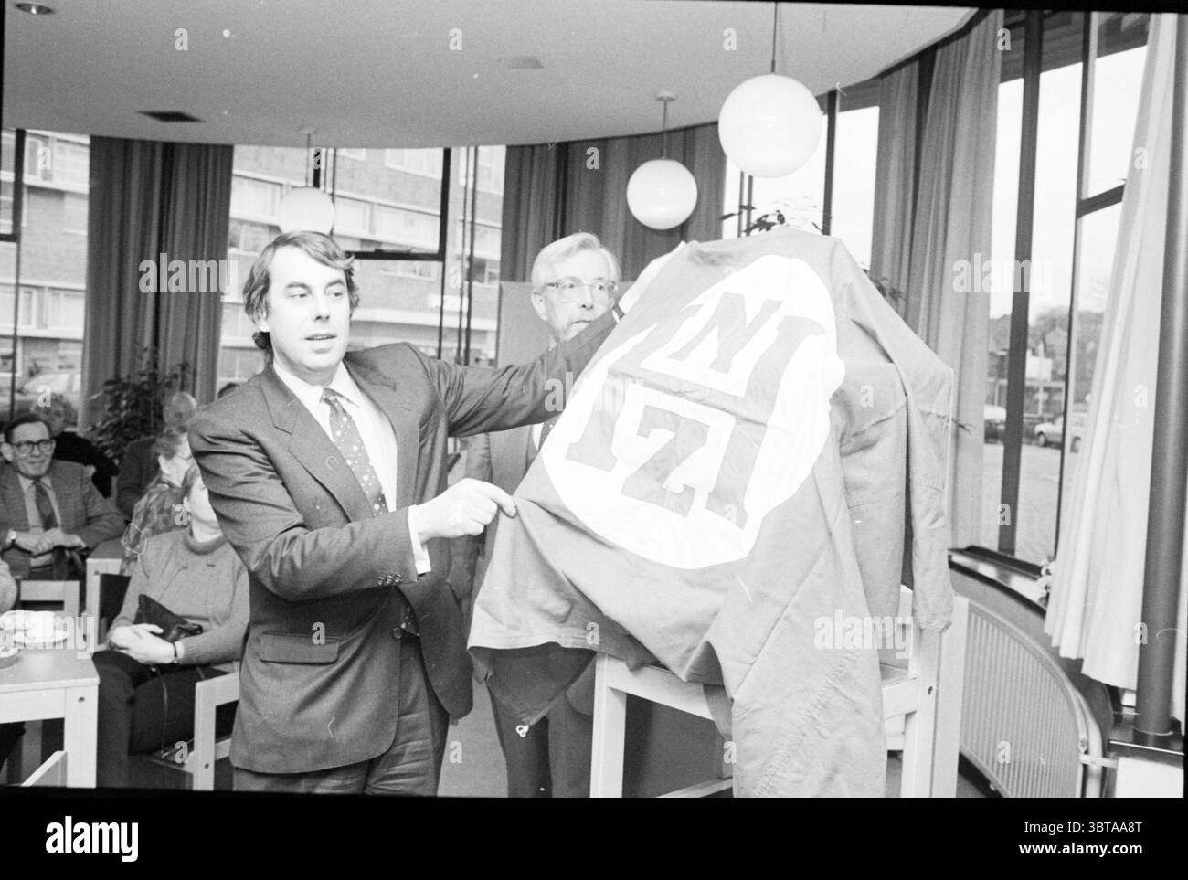 Inaugurazione della mostra a scartamento ridotto NZH Noord Zuid Hollandse Vervoersmaatschappij N.V. apertura aperture messa in servizio, Whizgle News, Dutch Desk, Paesi Bassi, 1950 - 2000 on 28-01-1988. Questi sono gli elementi dell'immagine. In un ambiente interno caldo e illuminato, un gruppo di persone è riunito in modo informale ma attento. La sala sembra essere un centro comunitario o uno spazio per riunioni, caratterizzato da ampie finestre che permettono alla luce naturale di filtrare, con vista sul verde esterno. Le pareti sono adornate da tende dai colori neutri che cadono a cascata, ammorbidendo la luce e contribuendo Foto Stock