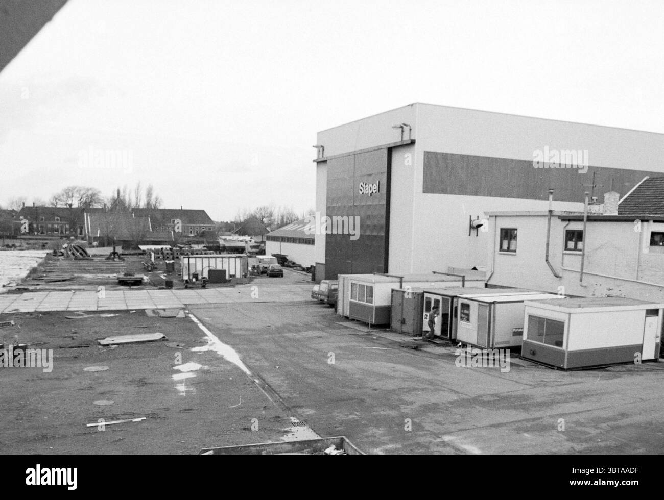 Panoramica del cantiere navale Stapel Spaarndam Shipping Spaarndam, Whizgle News, Dutch Desk, Paesi Bassi, 1950 - 2000 il 18-12-1986. Questi argomenti sono mostrati nell'immagine. La scena presenta un paesaggio industriale aspro, caratterizzato da un senso di abbandono e desolazione. In primo piano si estende un'ampia area lastricata, la sua superficie segnata da macchie di umidità e macchie di sporco, che suggeriscono una mancanza di manutenzione. Alcuni contenitori sparsi e strutture improvvisate sono posizionati in modo non uniforme, alcuni sembrano usurati e intemprati. Sulla destra si erge un grande e imponente edificio, il suo fa Foto Stock
