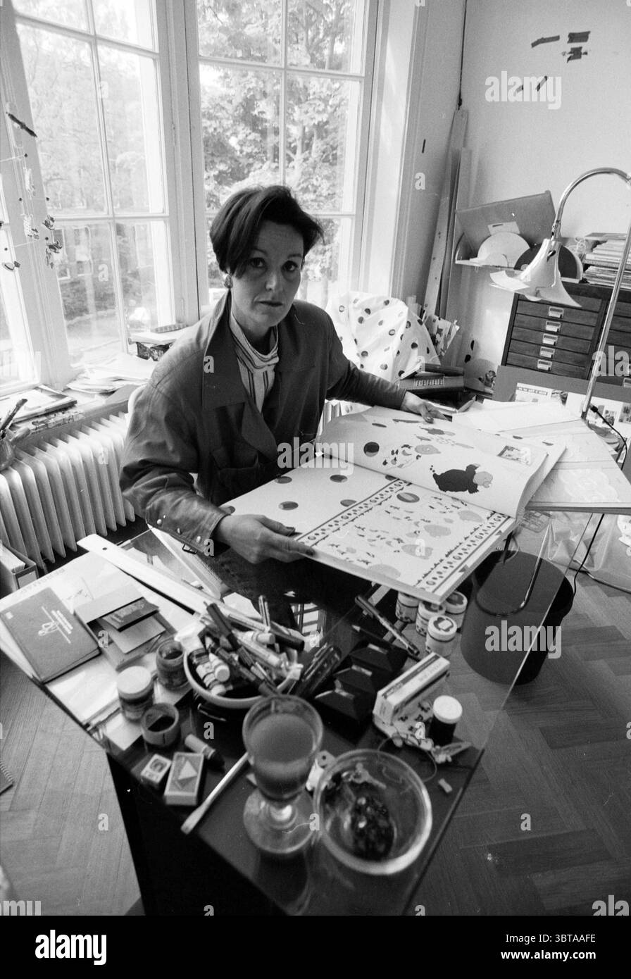 Sig.ra Mara IJsbouts designer, Whizgle News, Dutch Desk, Paesi Bassi, 1950 - 2000 il 22-05-1991. Questi sono gli elementi dell'immagine. La scena cattura uno spazio di lavoro pieno di energia creativa, illuminato dalla luce naturale che scorre attraverso le grandi finestre. Una donna siede su una scrivania disordinata, circondata da una vasta gamma di oggetti d'arte. La sua espressione è focalizzata, suggerendo una profonda concentrazione mentre esamina un foglio pieno di forme e motivi colorati. La scrivania è una miscela caotica di materiali: Tubi di vernice, pennelli e bottiglie di vari colori sono sparsi sulla sua superficie, accanto a sh Foto Stock