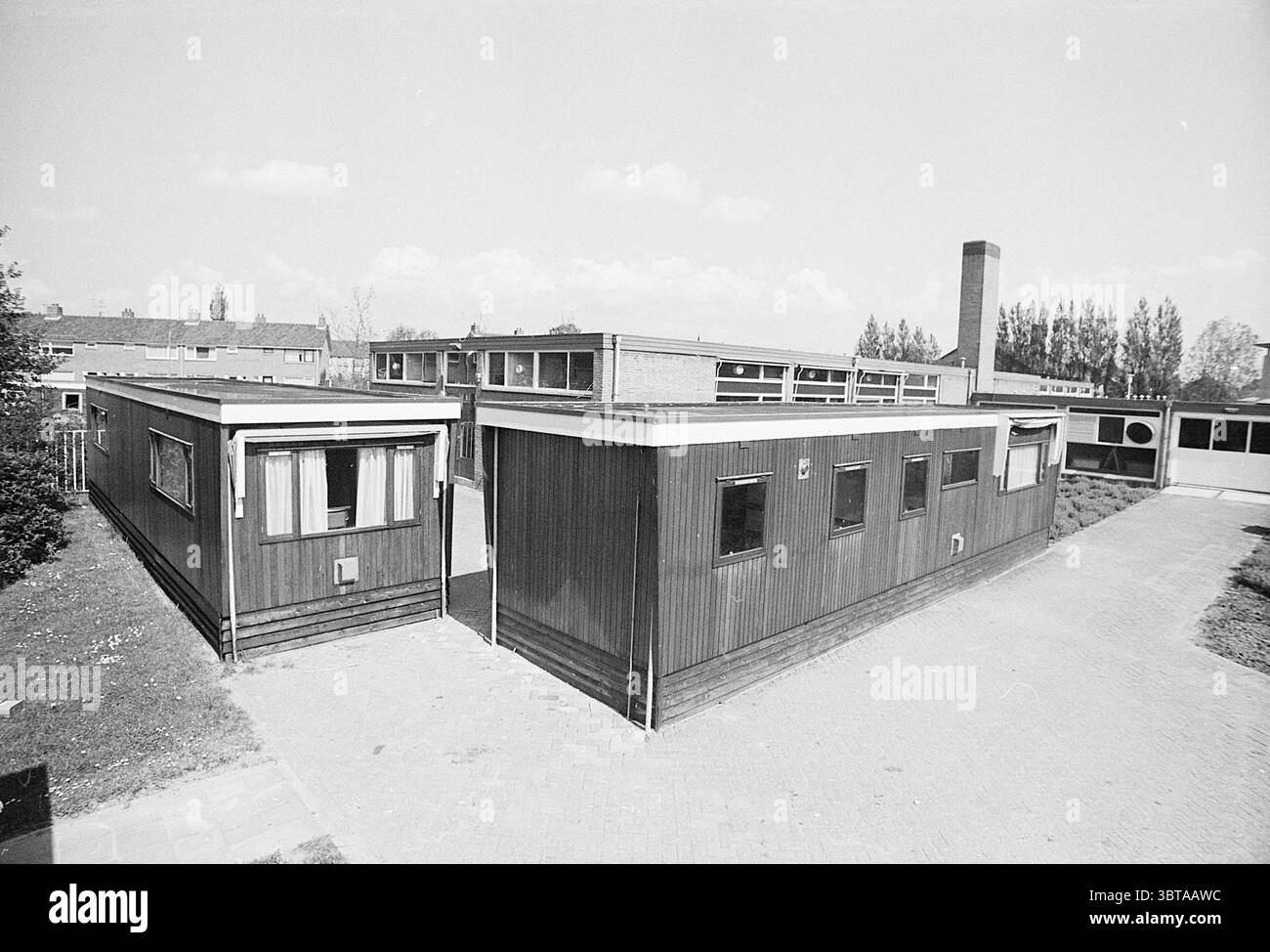 soc. Caravan est. Servizio esterno, Whizgle News, Dutch Desk, Paesi Bassi, 1950 - 2000 su 11-05-1972. Questi sono gli elementi dell'immagine. La scena raffigura una collezione di bassi edifici disposti in un layout angolare, caratterizzato da uno stile architettonico semplice ma funzionale. Le strutture sono caratterizzate da pannelli in legno che appaiono intemprati, con una tavolozza di colori tenui dominati da marroni e grigi, che donano un fascino rustico. Intorno agli edifici, un sentiero pavimentato si snoda attraverso macchie di erba. Il sentiero è fiancheggiato da un paesaggio sovrastato, suggerendo che la natura si riprende dolcemente Foto Stock