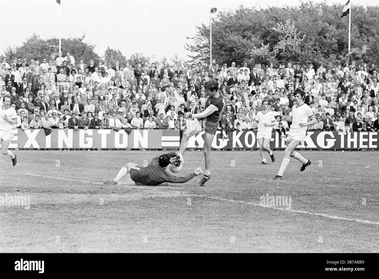 Match SC Telstar-HFC Haarlem Velsen., Whizgle News, Dutch Desk, Paesi Bassi, 1950 - 2000. Questi sono gli elementi dell'immagine. La scena cattura un momento dinamico su un campo di calcio durante una partita competitiva. In primo piano, un giocatore con un'uniforme scura sta tentando abilmente di calciare la palla, mentre un altro giocatore, vestito con un kit più leggero, si sta tuffando a terra, cercando di intercettare la palla. L'espressione sul volto del portiere trasmette determinazione, mettendo in evidenza la tensione del gioco. Lo sfondo è pieno di una grande folla di spettatori, molti dei quali sono vestiti in un mix di Foto Stock