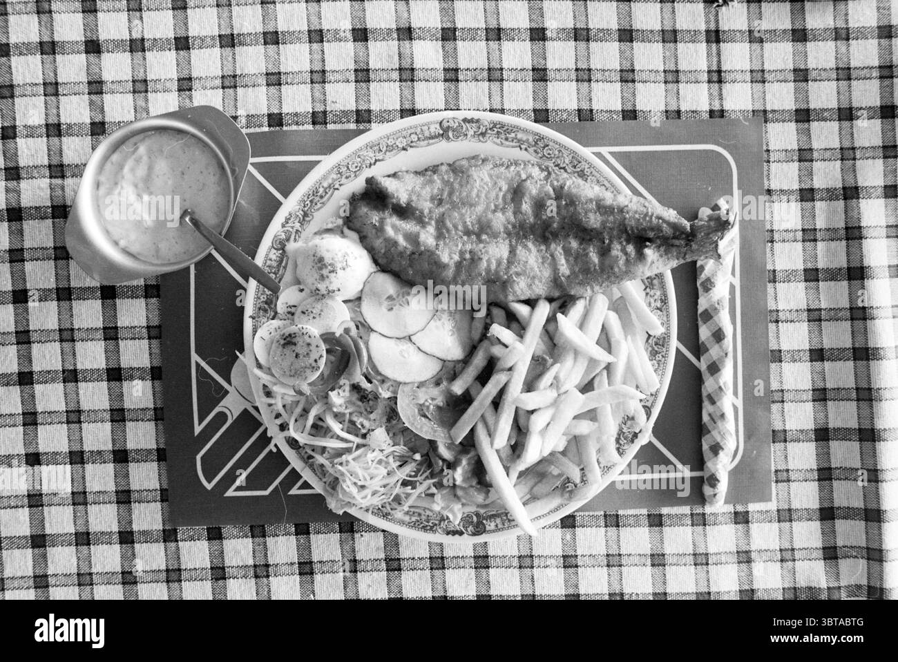 Piatto con pesce fritto pesca e altri pescivendoli pesce, Whizgle News, Dutch Desk, Paesi Bassi, 1950 - 2000 il 14-08-1989. Questi sono gli elementi dell'immagine. La scena mostra un pasto organizzato su una tovaglia a motivi caratterizzati da un classico design a scacchi. Al centro, un grande pesce fritto dorato viene presentato intero, la sua pelle croccante luccicante che mostra squame intricate. Intorno al pesce ci sono vari contorni, tra cui una porzione di patatine fritte dorate croccanti e cotte al vapore, e un variopinto assortimento di verdure fresche. Queste verdure sono caratterizzate da un vivace Foto Stock