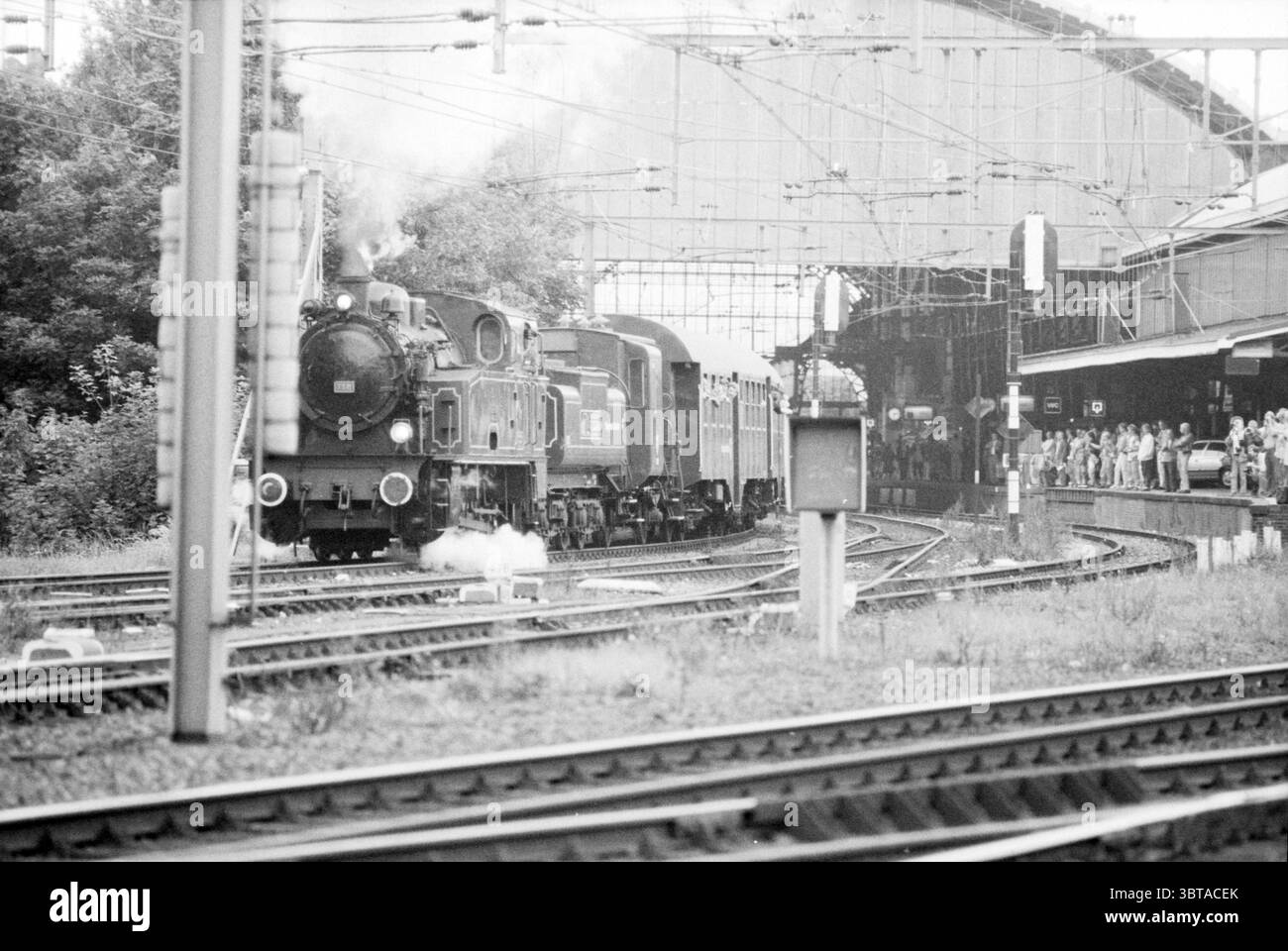 Open Day NS workshop 150 anni di NS Open Day e Open House Railways, Whizgle News, Dutch Desk, Paesi Bassi, 1950 - 2000 il 23-09-1989. L'immagine include questi argomenti. In una vivace stazione ferroviaria, una locomotiva a vapore d'epoca sprigiona bianchi colpi di vapore, creando un senso di nostalgia. La locomotiva è prevalentemente nera, adornata con accenti metallici che brillano leggermente sotto la luce ambientale. Tira una serie di carrozze intemprate, ognuna delle quali riflette sfumature di verde scuro e ruggine. La stazione stessa è dotata di un ampio baldacchino in alto che incornicia la scena, gettando una stufa d'ombra Foto Stock