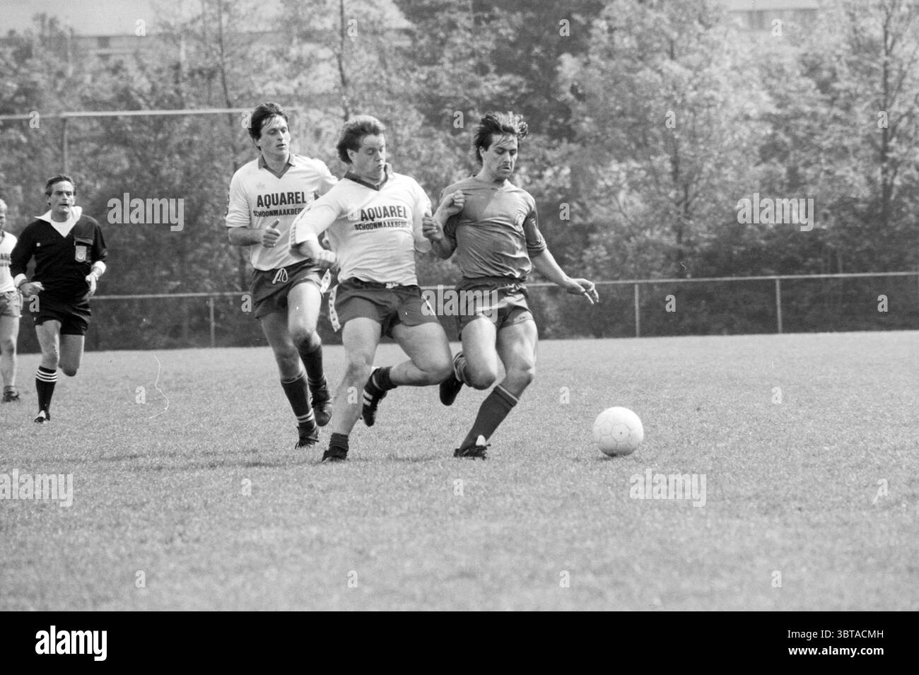 Partita di calcio Nieuw Vennep e campioni, Whizgle News, Dutch Desk, Paesi Bassi, 1950 - 2000 il 05-1987. L'immagine contiene questi argomenti. La scena è ambientata su un campo da calcio erboso, vivace e vivace. Due giocatori, entrambi con maglie bianche con logo "AQUAREL" impresso in grassetto sul davanti, stanno inseguendo da vicino un avversario che indossa una maglia rossa brillante. L'azione è incentrata sul giocatore in rosso, che sta guidando abilmente la palla lungo il campo, il suo corpo angolato in uno sprint dinamico. L'erba è un verde vivace, punteggiato da toni della terra del recente spettacolo teatrale, e il sett Foto Stock