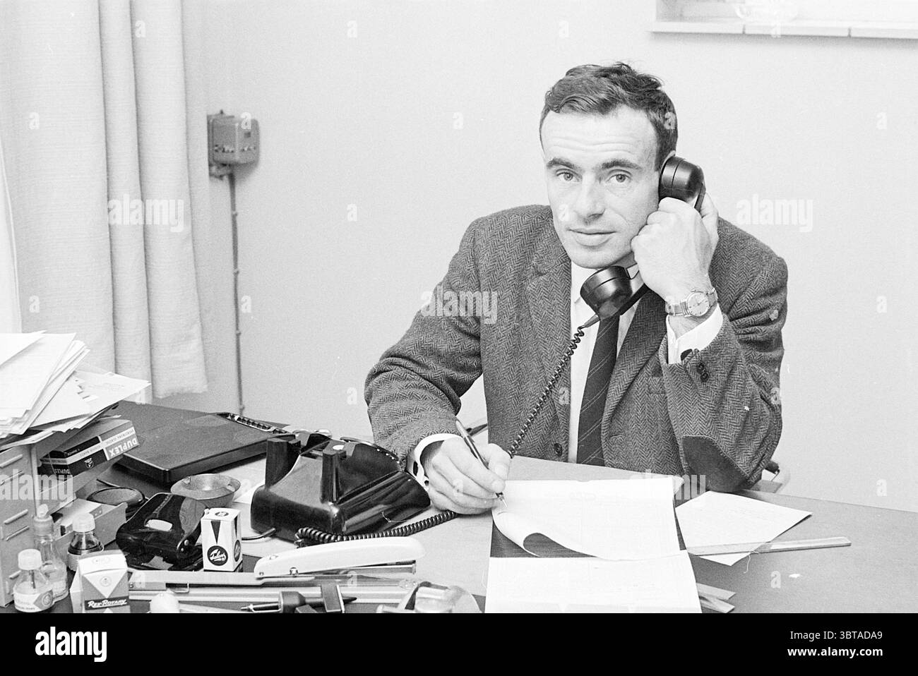 Ritratto di Koekendijk Portraits, Whizgle News, Dutch Desk, Paesi Bassi, 1950 - 2000 il 21-05-1969. L'immagine mostra questi argomenti. Un uomo ben vestito siede a una scrivania, impegnato in una conversazione telefonica. Ha i capelli corti e ordinati e sembra concentrato, con un leggero sorriso che suggerisce di essere in una piacevole conversazione. Il suo abbigliamento include un abito scuro, una camicia bianca e una cravatta, che conferisce un'aria di professionalità. Lo spazio di lavoro intorno a lui è ingombrante ma organizzato, con diversi fogli di carta sparsi per la scrivania. Una pila di file si trova su un lato, accanto a un telefono d'epoca, W Foto Stock