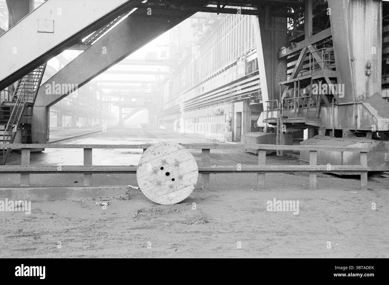 Altiforni - foto archivio Blast Fornace, Whizgle News, Dutch Desk, Paesi Bassi, 1950 - 2000 il 18-01-1982. L'immagine contiene questi argomenti. La scena cattura un ampio ambiente industriale, con un ampio spazio aperto dominato da macchinari pesanti ed elementi strutturali. Le imponenti cornici e travi metalliche creano un effetto labirintico, riempiendo lo sfondo di un senso di scala e complessità. Le superfici sono molto ruvide, con segni visibili di usura e polvere, a indicare una lunga storia di utilizzo. In primo piano, una grande bobina di legno si trova abbandonata su un terreno polveroso, esso Foto Stock