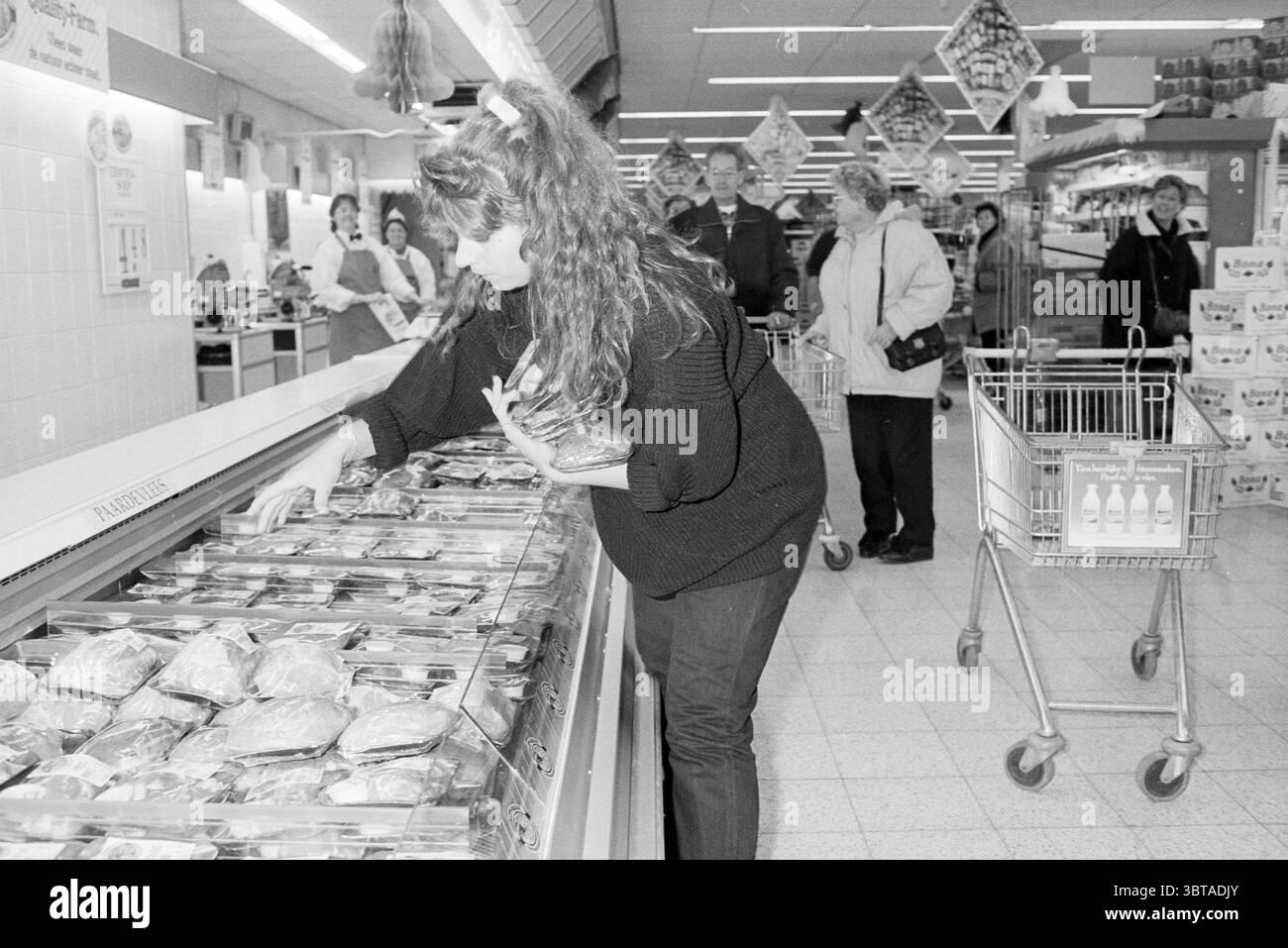 Vomar IJmuiden a tre minuti dai negozi di IJmuiden Paesi Bassi, Whizgle News, Dutch Desk, Paesi Bassi, 1950 - 2000 dal 13-12-1993. Questi argomenti vengono visualizzati nell'immagine. In un vivace negozio di alimentari, la scena è piena di diversi acquirenti impegnati nelle loro attività. Una donna con voluminosi capelli ricci, vestita con un maglione nero, si appoggia su una vetrina aperta piena di carni confezionate. La sua espressione concentrata suggerisce che sta selezionando attentamente i suoi articoli. Nelle vicinanze, un paio di persone anziane si trovano in una tranquilla conversazione, prendendo in considerazione la vasta gamma di prodotti disponibili. Sembrano confortabili Foto Stock