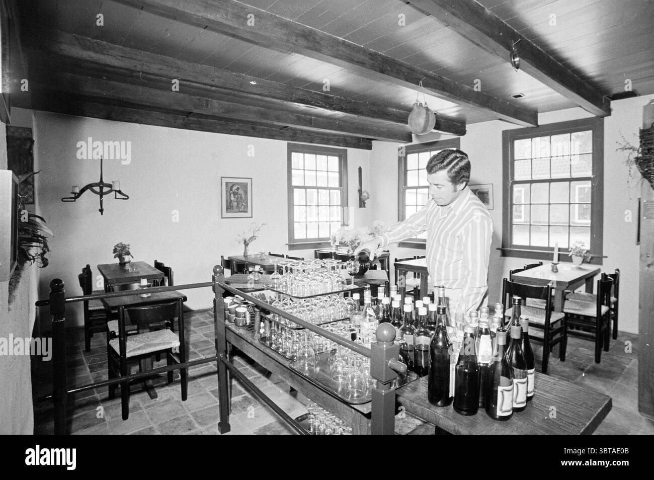 Interni di Het Roode Hert Interior, Whizgle News, Dutch Desk, Paesi Bassi, 1950 - 2000 il 09-06-1973. L'immagine contiene questi argomenti. La scena raffigura un interno accogliente e invitante di un ristorante o bar, caratterizzato da un fascino rustico. Travi in legno abbelliscono il soffitto, esaltando il calore dell'atmosfera. Le pareti sono dipinte in toni delicati e neutri, creando uno sfondo rilassante. In primo piano, un uomo è impegnato a versare una bevanda da una bottiglia, concentrandosi intensamente sul suo compito. Indossa una camicia a righe, aggiungendo un tocco di stile al suo aspetto. L'area del bar e' fiancheggiata da glasswa Foto Stock
