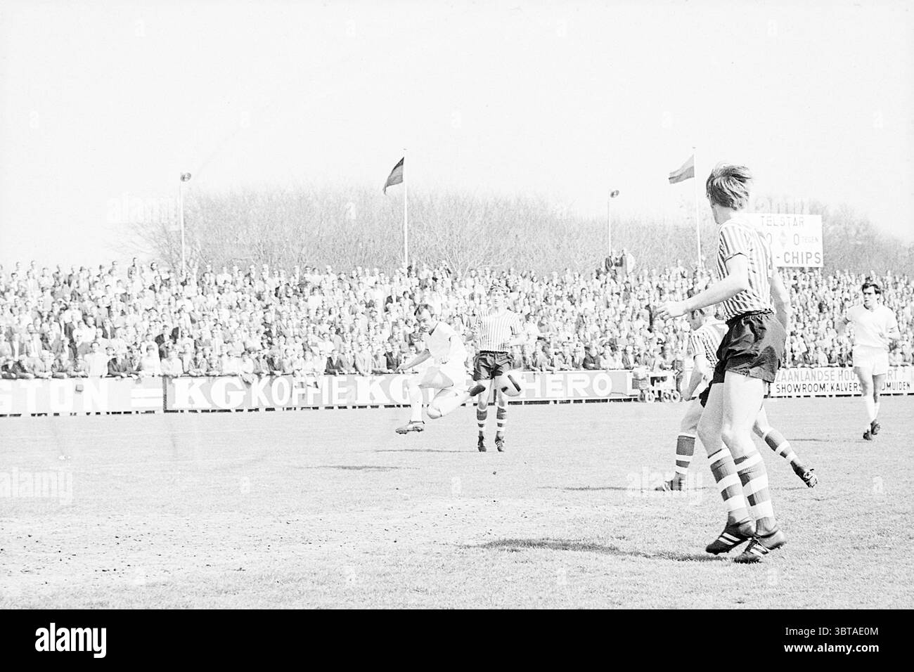 Telstar Football Match., Whizgle News, Dutch Desk, Paesi Bassi, 1950 - 2000. L'immagine mostra questi argomenti. La scena raffigura una vivace partita di calcio all'aperto, sullo sfondo di una grande folla riunita in una giornata di sole. I tifosi sono densamente affollati di tribune e la loro attenzione è concentrata sul gioco, con espressioni diverse che vanno dall'eccitazione alla tensione. In primo piano, due giocatori sono impegnati nell'azione; uno è in movimento, navigando abilmente sul campo e preparandosi a passare la palla, mentre l'altro è pronto, pronto per il gioco successivo. Gli atleti noi Foto Stock